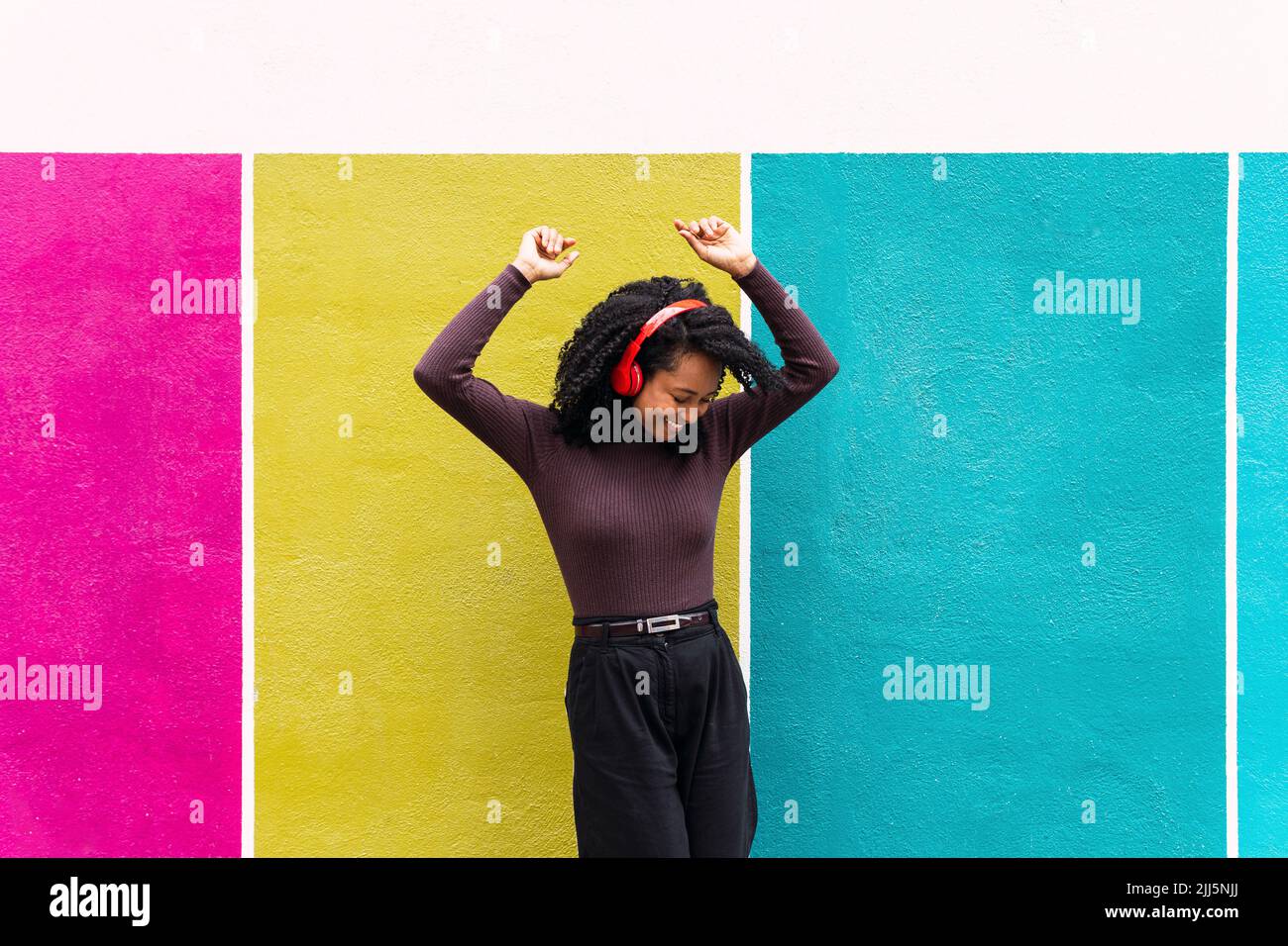 Fröhliche Frau, die mit erhobenen Armen tanzt und Musik über kabellose Kopfhörer vor einer mehrfarbigen Wand genießt Stockfoto