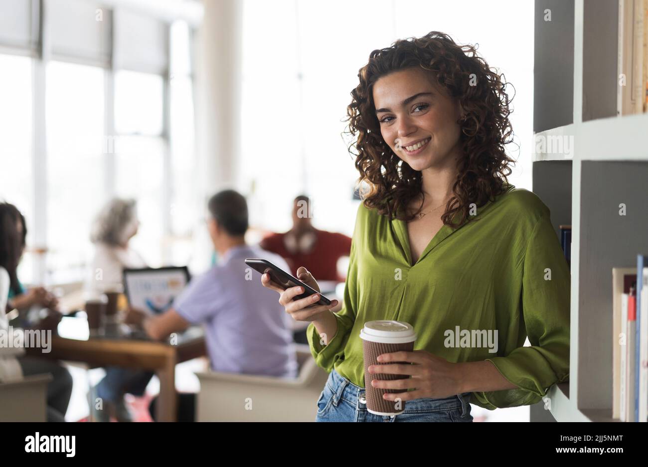 Lächelnde Geschäftsfrau mit Smartphone und Einwegbecher, die im Büro auf dem Regal gelehnt ist Stockfoto