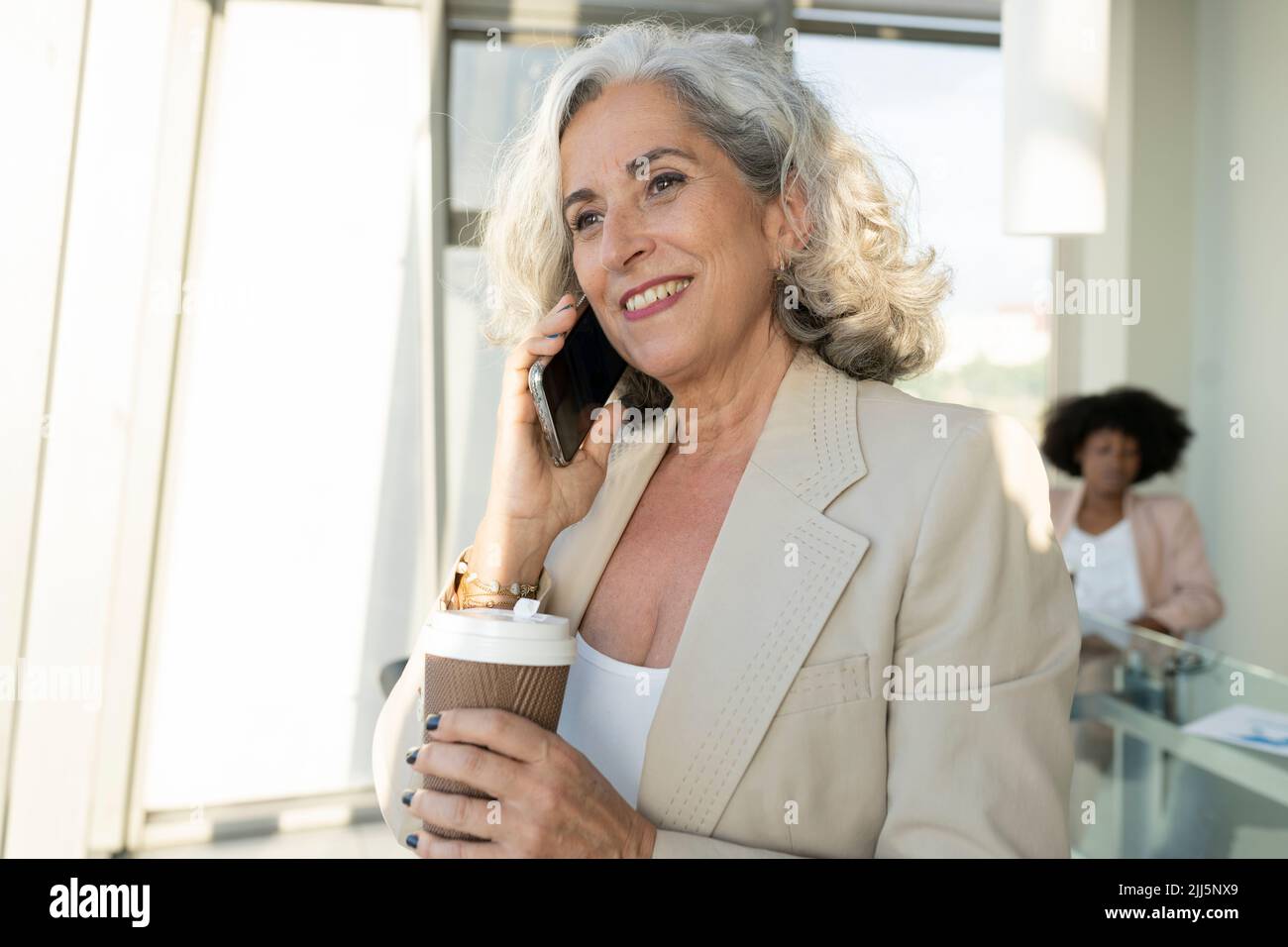 Leitende Geschäftsfrau mit einem Einwegbecher, der im Büro auf einem Smartphone spricht Stockfoto