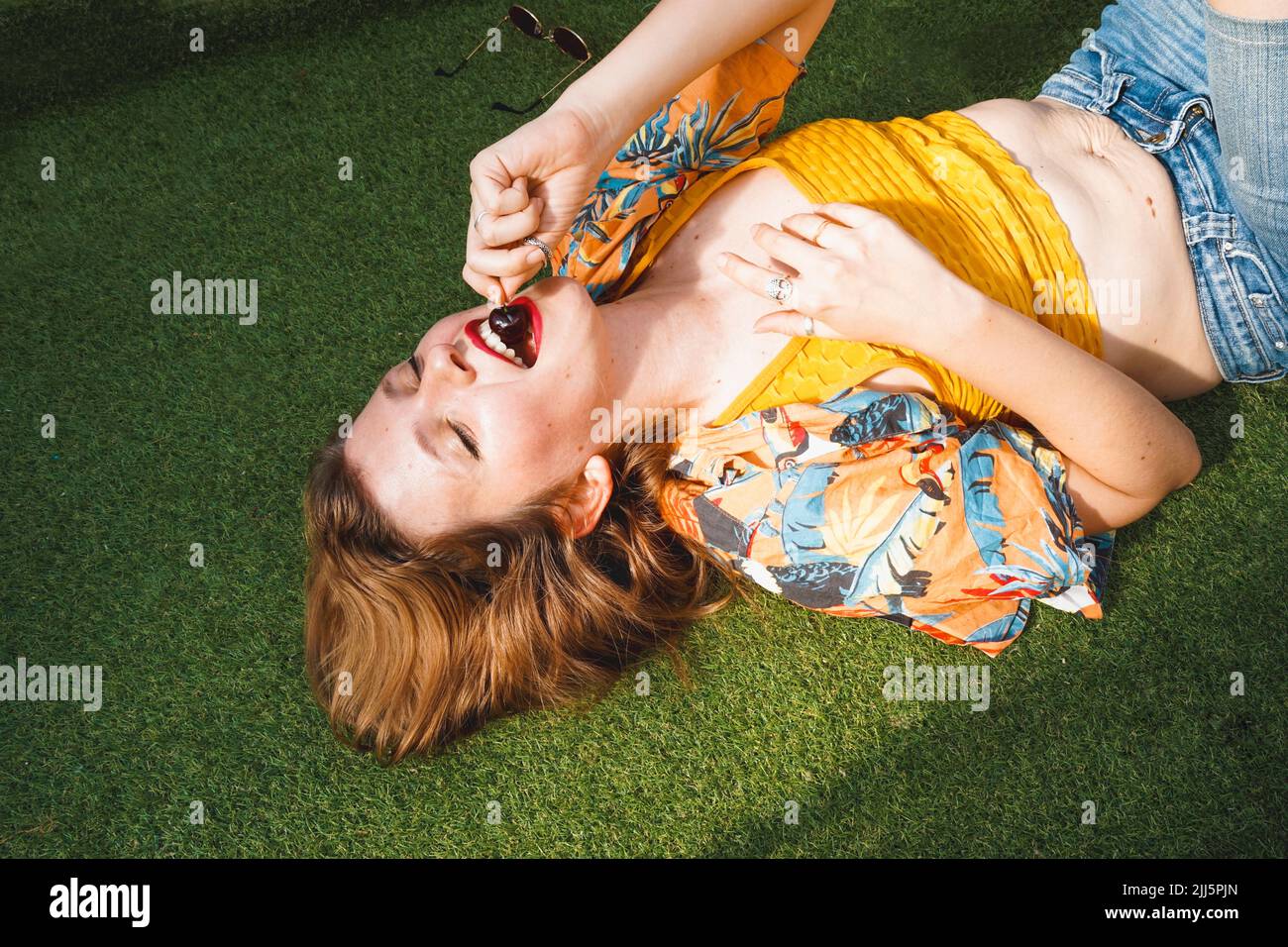 Glückliche Frau mit geschlossenen Augen, die Kirsche isst und auf Gras liegt Stockfoto