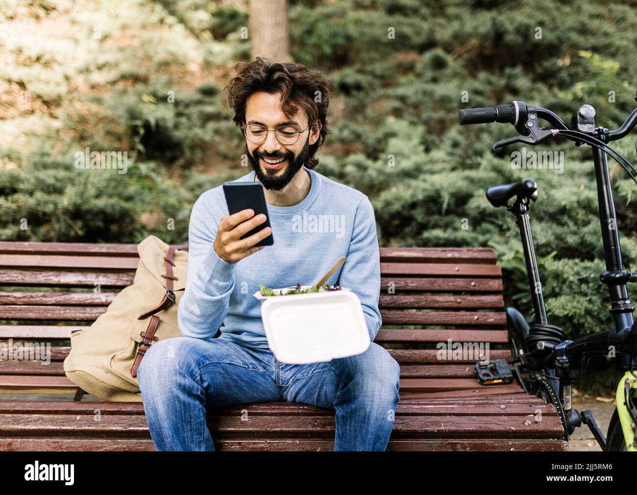 Lächelnder Mann mit Lunchbox am Telefon auf der Bank sitzend Stockfoto