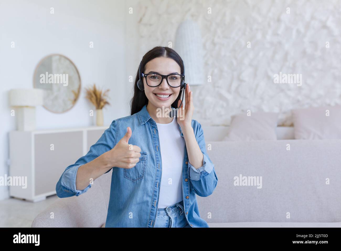 Schöne Frau, die zu Hause mit einem Headset für Videoanruf arbeitet und studiert, lächelt und schaut auf die Kamera, hält den Finger hoch, sitzt auf dem Sofa im Wohnzimmer Stockfoto