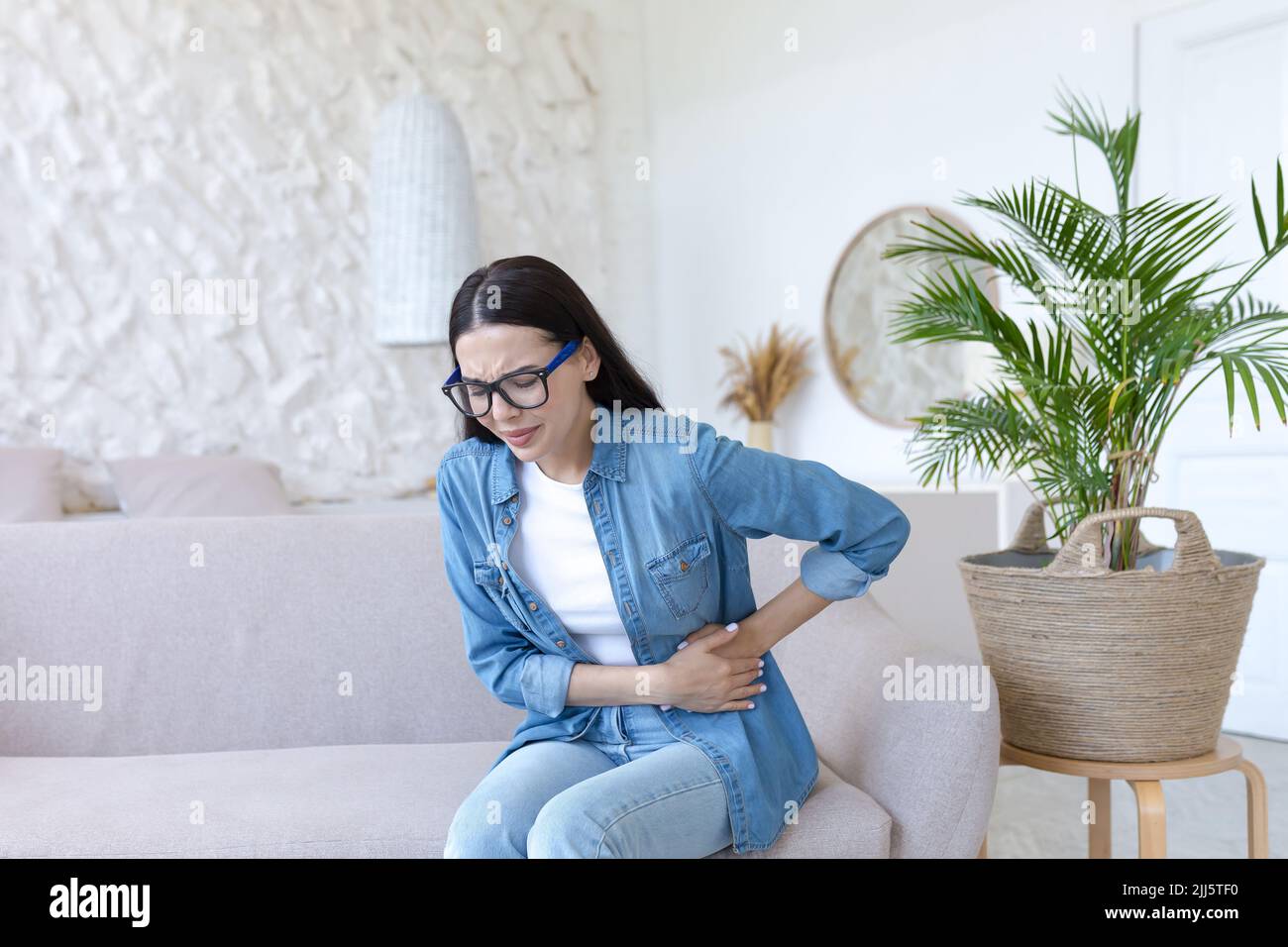 Eine junge, schöne Frau sitzt zu Hause auf der Couch, krank, hat starke Schmerzen in Magen und Seite Stockfoto