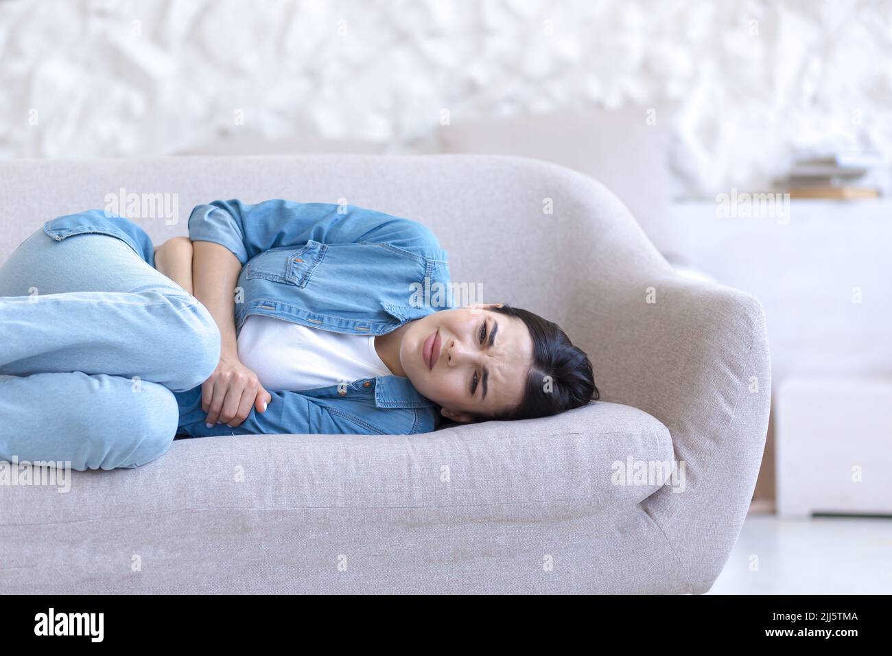 Junge, schöne Frau allein zu Hause, auf dem Sofa liegend, Nahaufnahme, verärgert, mit schweren Magenschmerzen Stockfoto