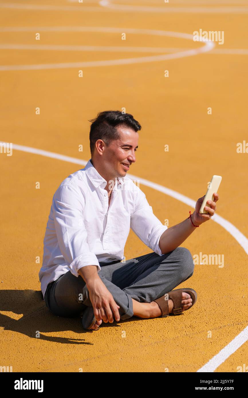 Lächelnder Mann, der mit gekreuzten Beinen auf dem Sportplatz mit dem Mobiltelefon sitzt Stockfoto
