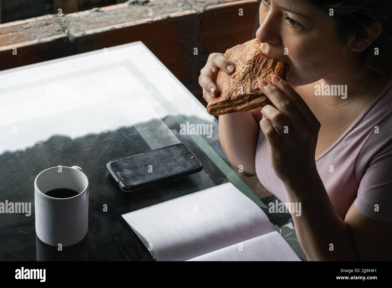 Wunderschöne Latina-Frau, die vor dem College ein Sandwich zum Frühstück aß, Kaffee in einem weißen Becher trank und ihr Handy neben einem BL hielt Stockfoto