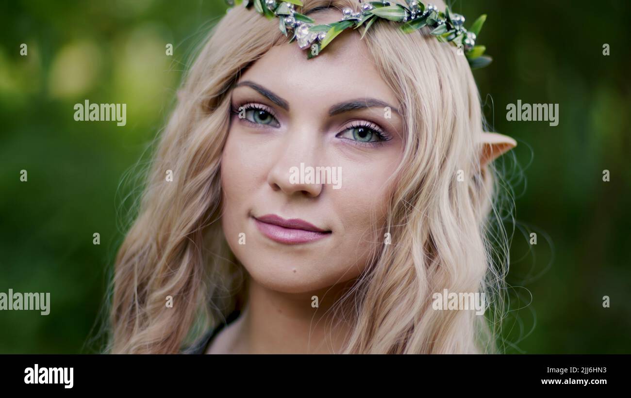 Porträt einer jungen Frau in Cosplay-Elf-Kleidung mit Make-up auf grünem Hintergrund. Fantastischer Look, langes blondes Haar, Waldkrone. Halloween-Konzept. Stockfoto