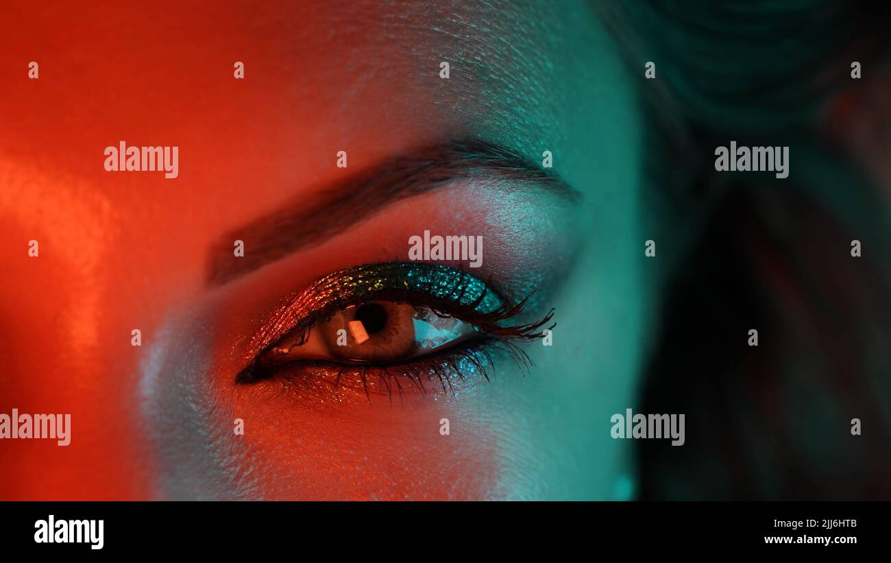 Nahaufnahme des weiblichen Auges unter orangeblauem Neonlicht. Weibchen mit schönem Make-up, Glitzer Schatten. Beauty, Kosmetik, Nightclub Lifestyle Konzept. Stockfoto