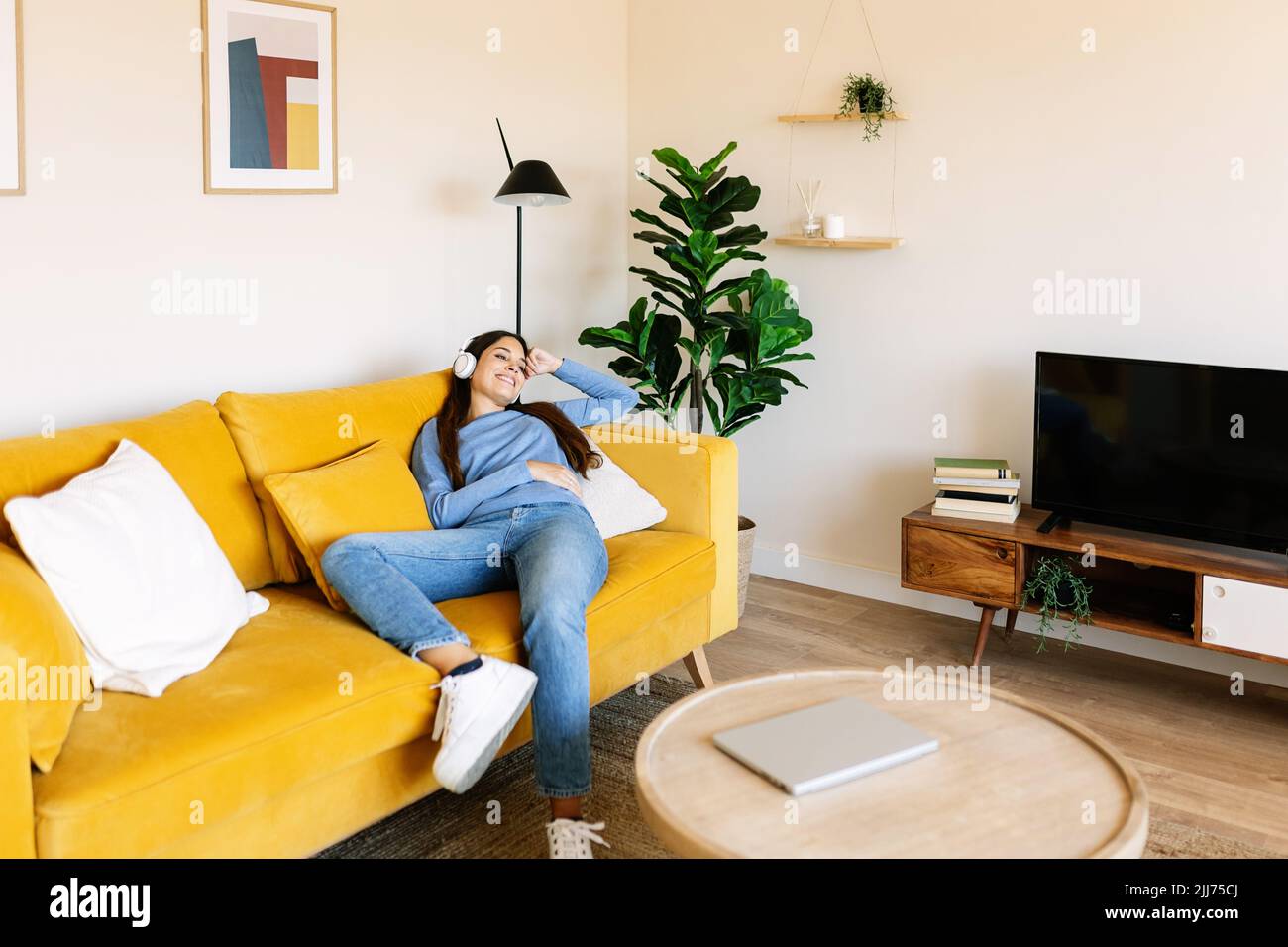 Entspannt lächelnde junge Frau sitzt auf dem Sofa, während sie zu Hause Musik hört Stockfoto
