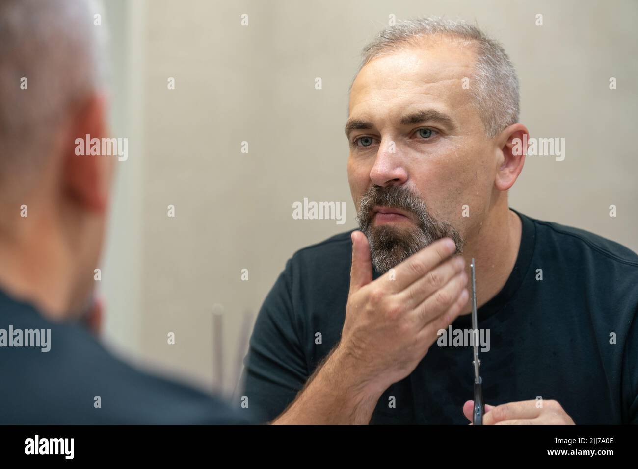 Ein gutaussehender Mann mittleren Alters, der mit einer Schere seinen Bart ein bisschen abschneidet und die Form fixiert Stockfoto