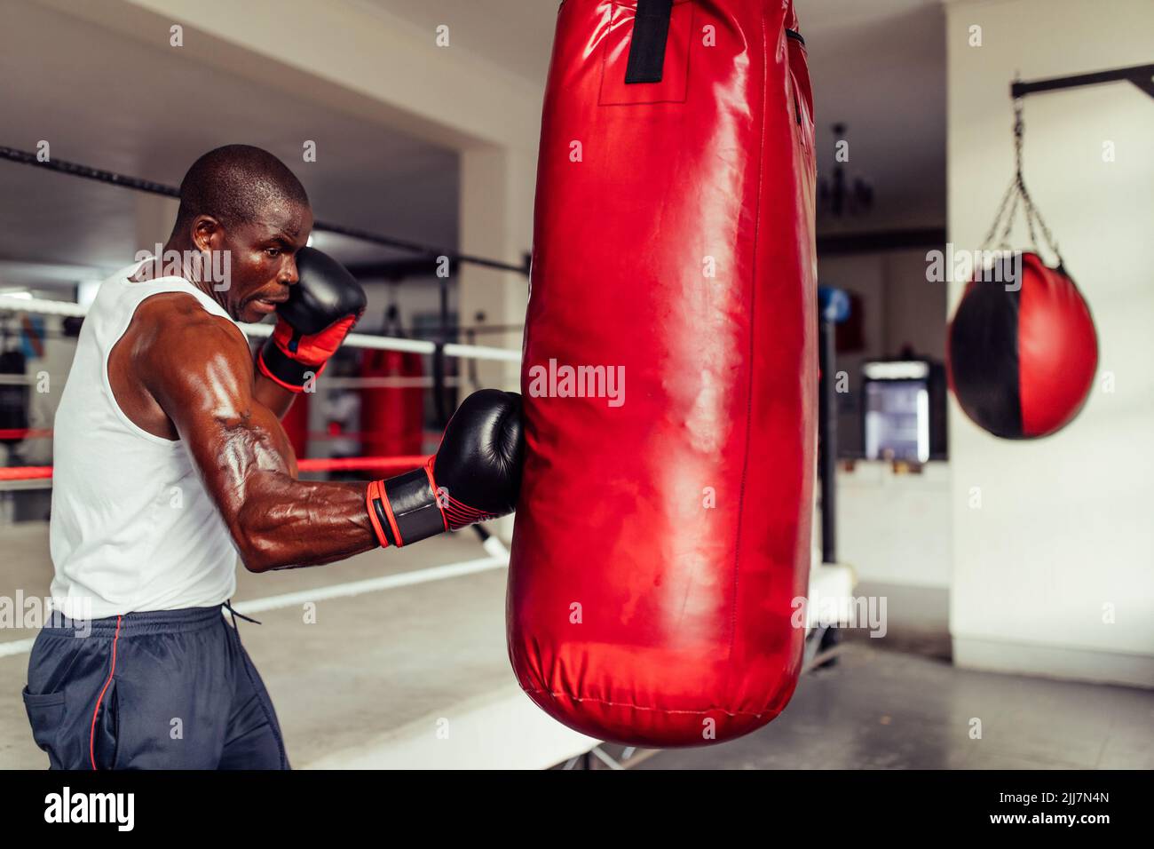 Muskulöser afrikanischer Boxer übt seine Schläge auf einem roten Boxsack aus Leder. Sport junger Mann, der eine Trainingseinheit in einer Boxhalle hat. Stockfoto
