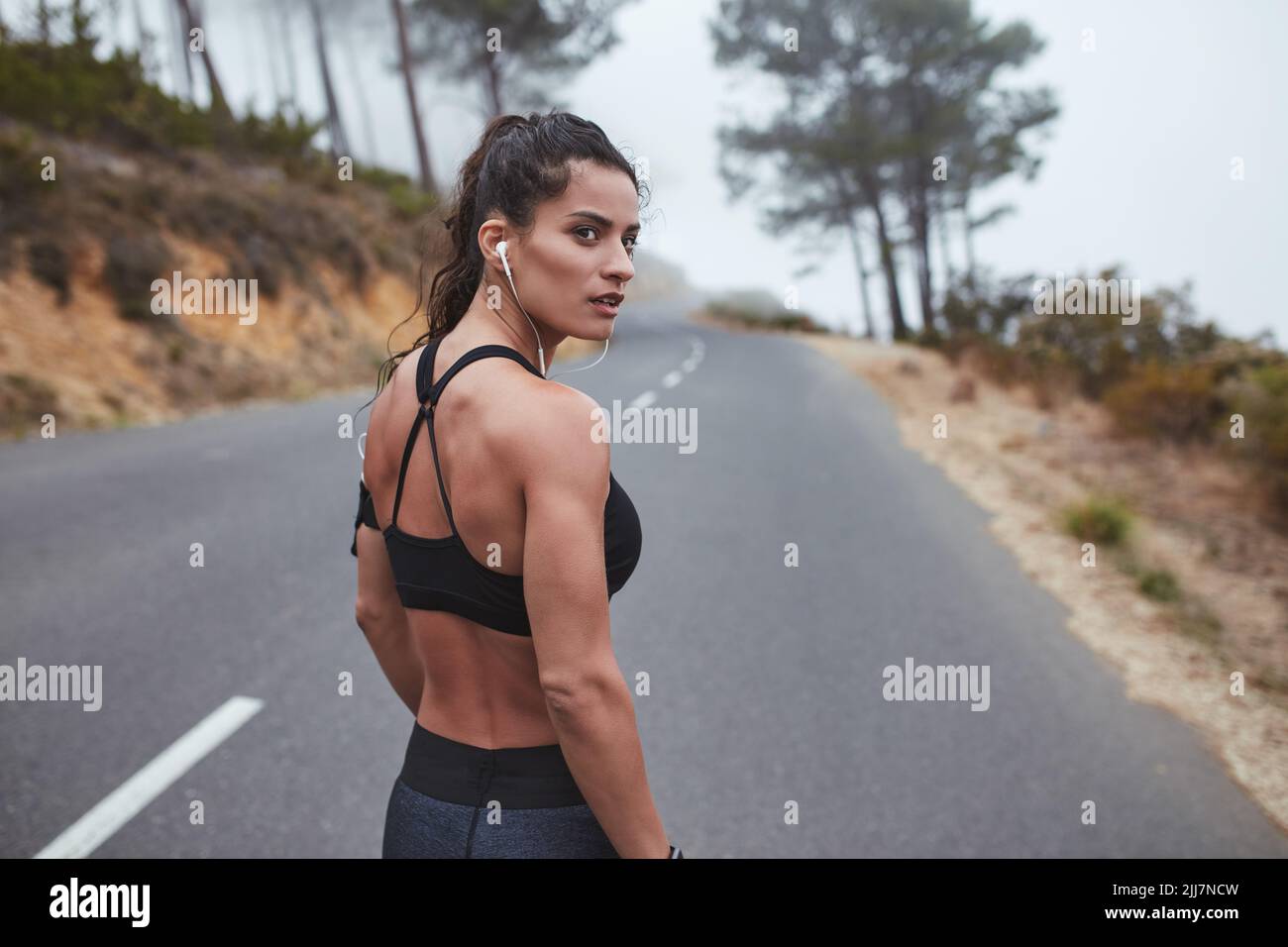 Muskulöse junge Frau, die wegschaut, während sie in Sportkleidung auf einer Straße steht. Läuferin, die ein morgendliches Training im Freien absolviert. Stockfoto