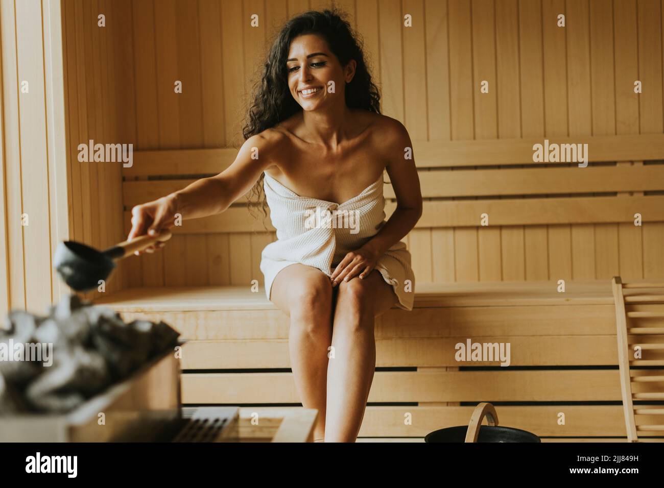 Attraktive junge Frau, die in der Sauna Wasser auf heißen Stein gießt Stockfoto
