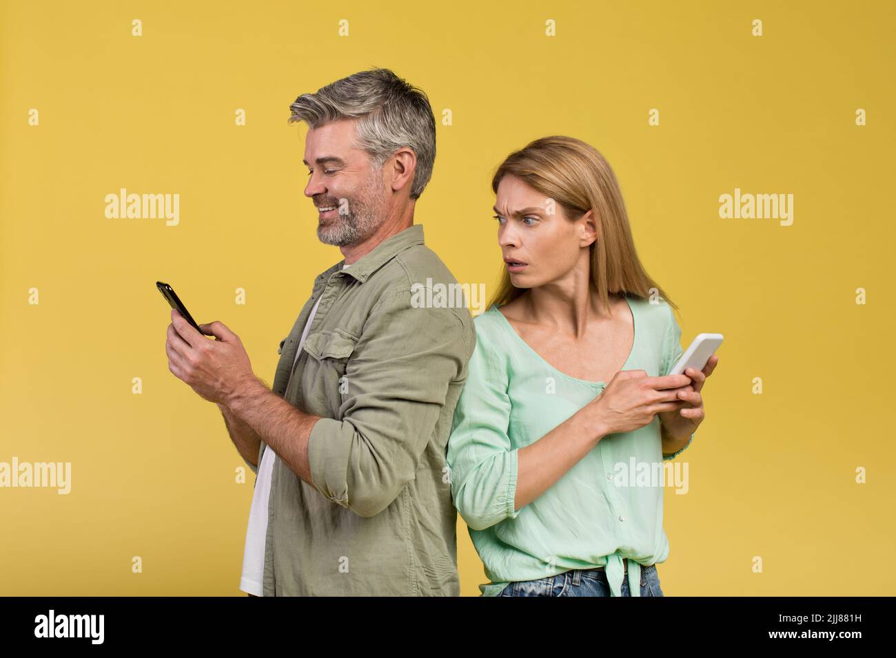 Schockierte Frau, die ihren glücklichen Mann mit dem Smartphone ausspioniert und sich über gelben Hintergrund zurück an den Rücken stellt Stockfoto