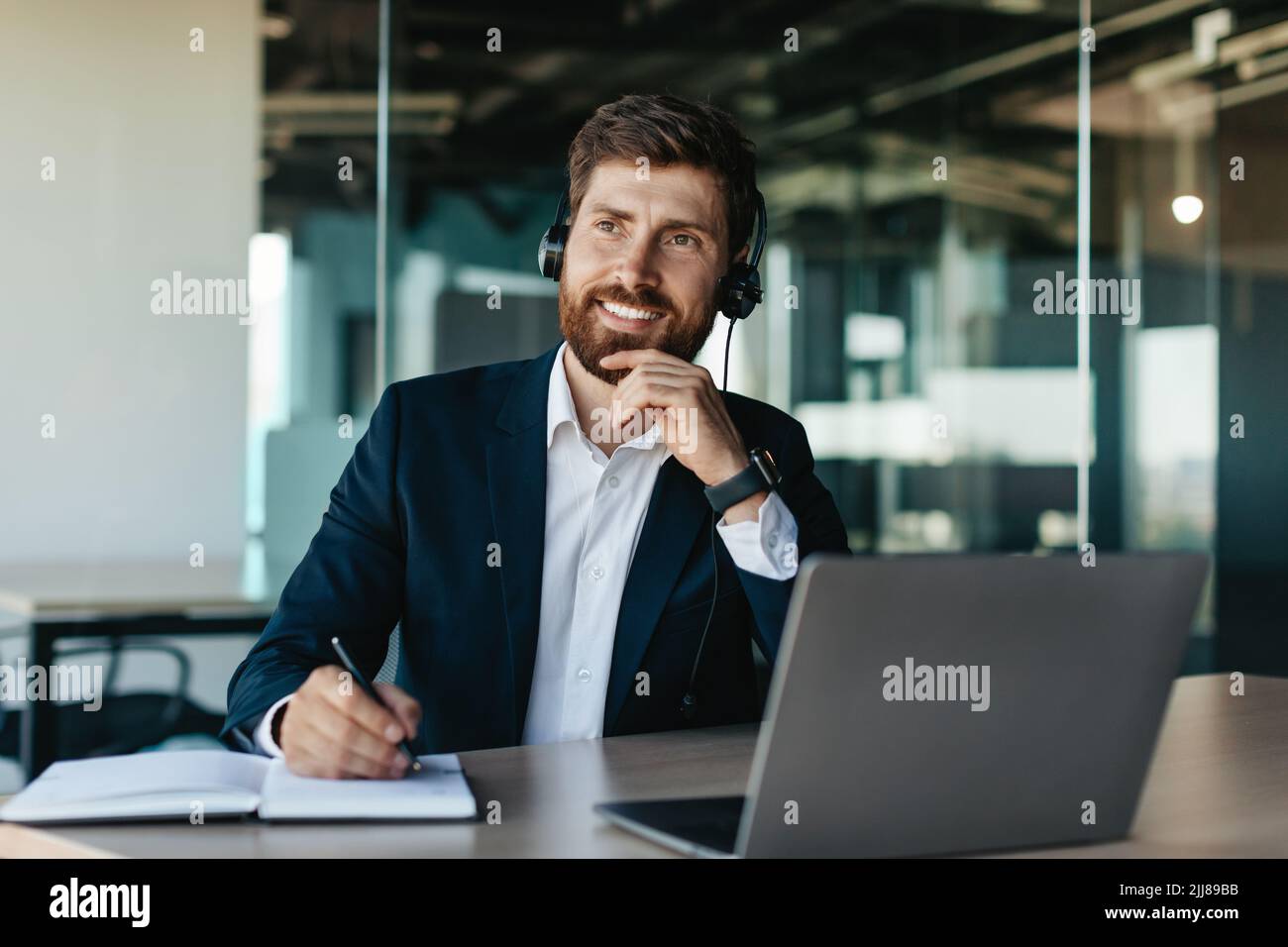 Ein glücklicher, nachdenklicher Geschäftsmann mit Headset, der am Laptop und im Notebook arbeitet und am Schreibtisch im Büroinneren sitzt Stockfoto