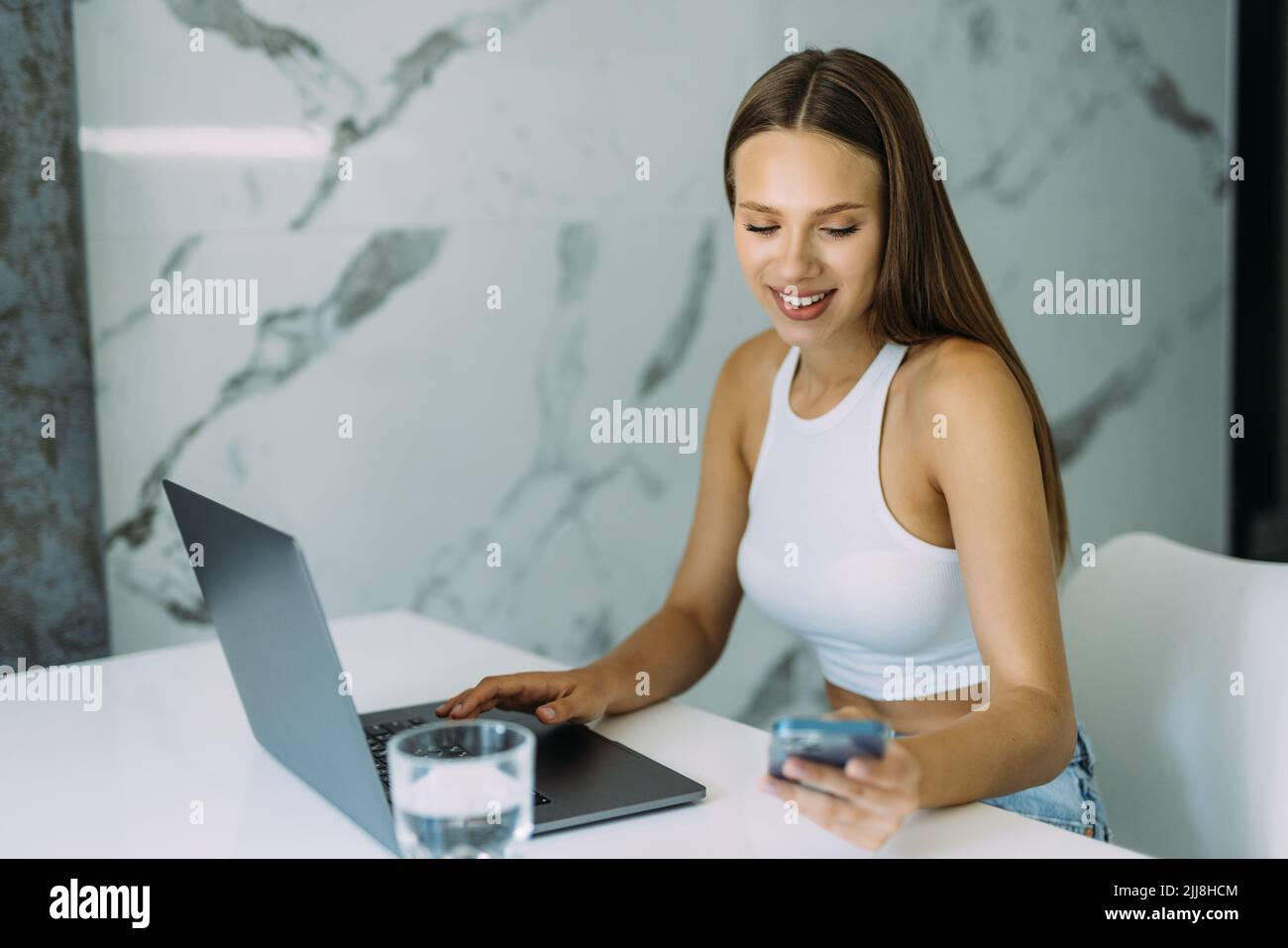 Selbstbewusste, freiberufliche Mitarbeiterin aus dem lateinamerikanischen ausland sitzt am Küchentisch und sucht Dateien auf dem Telefon, um sie über das drahtlose Heimnetzwerk auf dem Laptop weiterzuleiten. Schlau Sie Stockfoto
