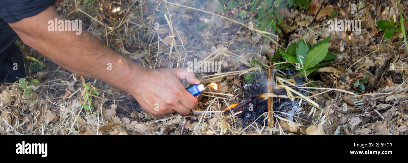 Bild der Hand eines Brandstifters, der mit einem Feuerzeug in einem Holz ein Feuer ausbricht. Hinweis auf das Problem der Waldbrände. Horizontales Banner Stockfoto