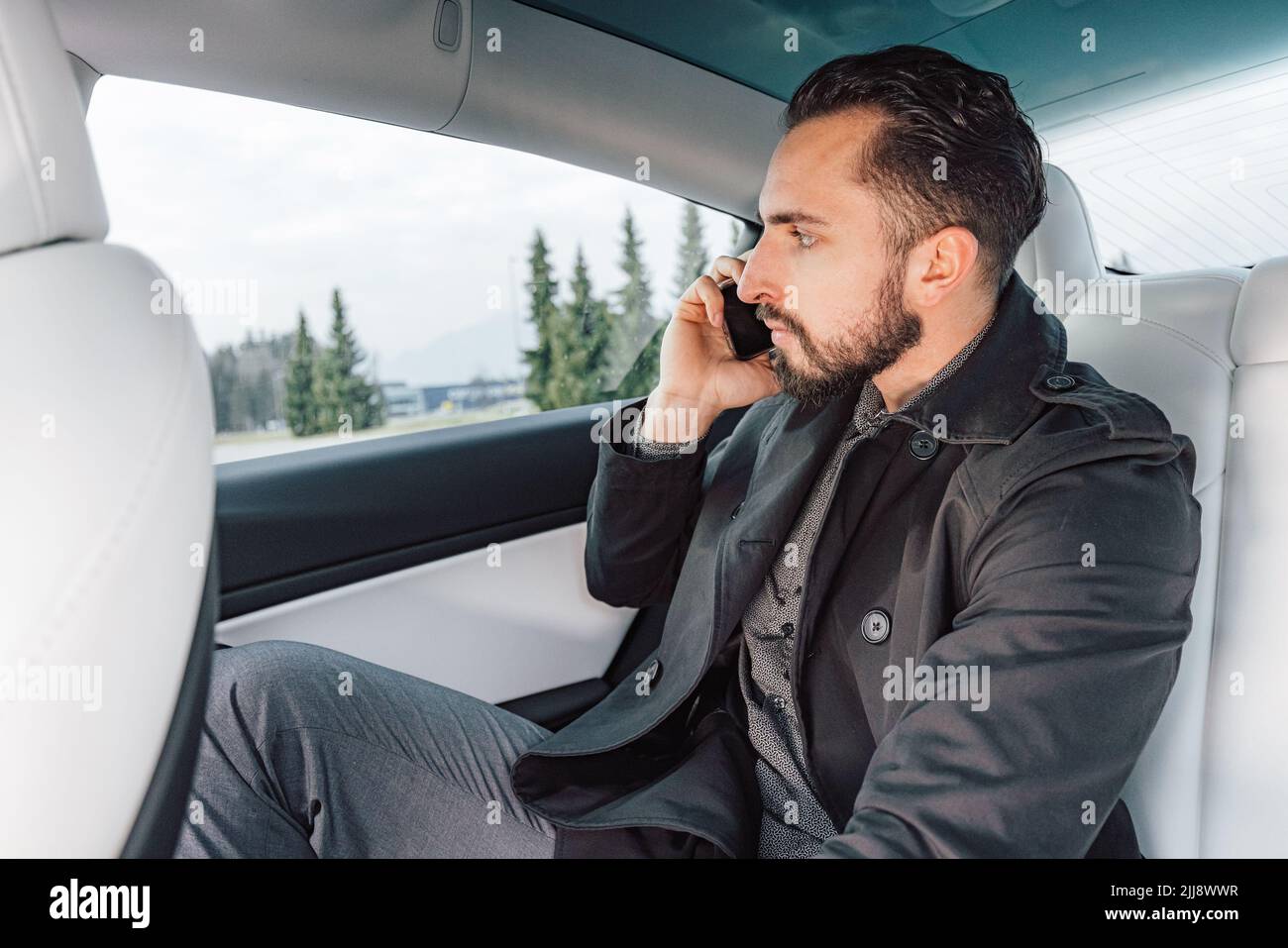Kaukasischer junger erfolgreicher Mann, der ernsthaft auf dem Smartphone spricht, während er auf dem Rücksitz eines fahrenden Autos sitzt Stockfoto