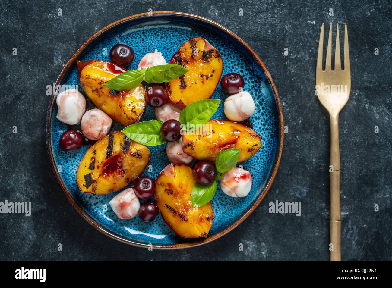 Gegrillter Pfirsichsalat mit Mozzarella, roter Kirsche, grünem Basilikum und Sauce. Frischer Gourmet-Salat in Teller auf schwarzem Hintergrund, Nahaufnahme, Draufsicht Stockfoto