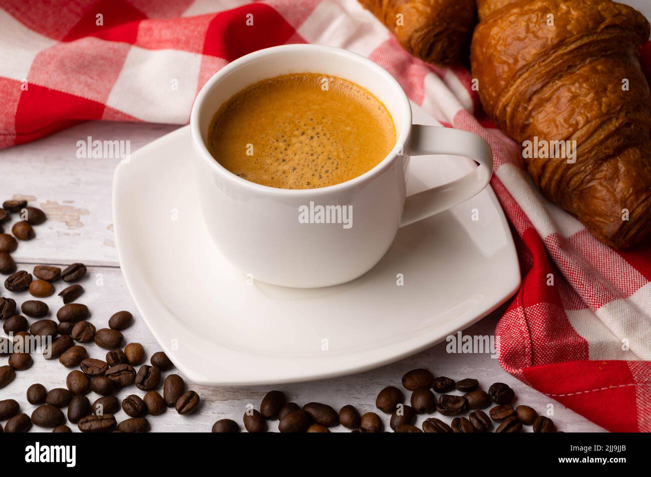 Eine Tasse Kaffee mit Croissants und verschütteten Kaffeebohnen auf einem weißen Holztisch. Um ein rot-weiß kariertes Küchentuch. Stockfoto