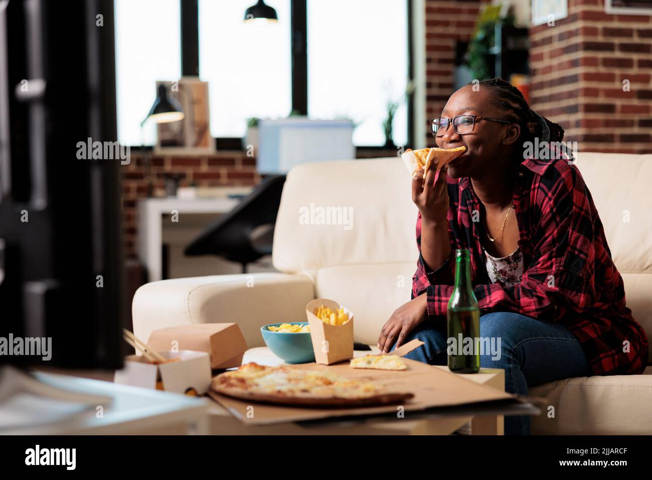 afroamerikanische Frau, die Pizza isst und sich einen Film ansieht, sich auf der Couch entspannt und Essen zum Mitnehmen geliefert. Genießen Sie Fast-Food-Mahlzeiten vom Takeout-Paket, um Filme im Fernsehen anzusehen. Stockfoto