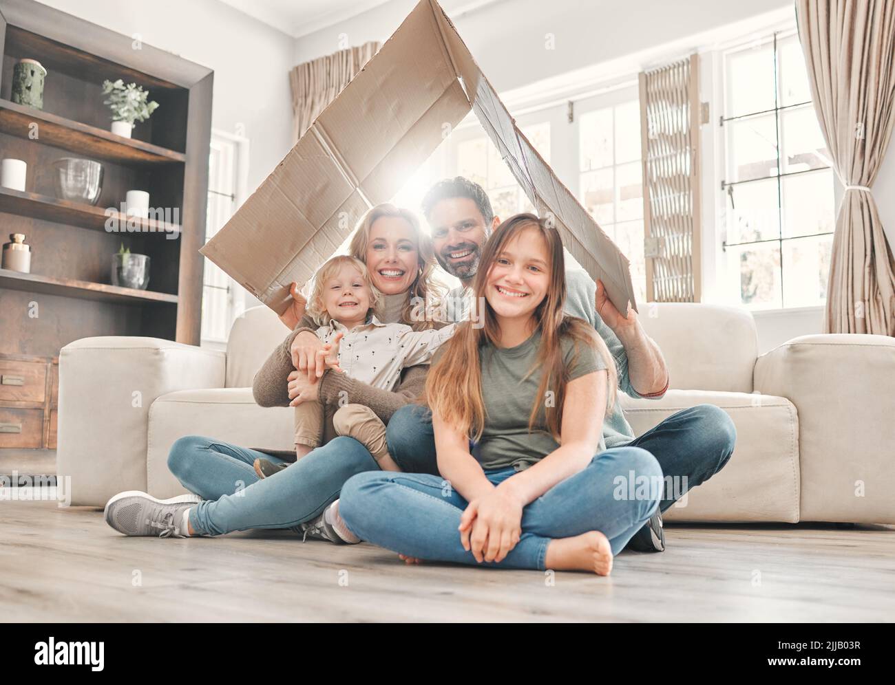 Die Familie ist es, was ein Haus zu einem Zuhause macht. Eine junge Familie hält einen Pappkarton in der Hand, um zu Hause ein Dach zu bilden. Stockfoto