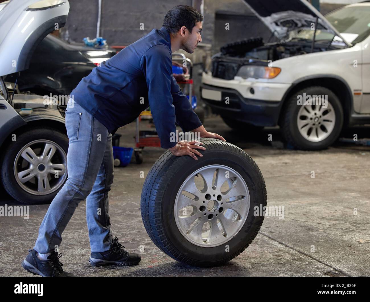 Reifenwechsel. Ganzkörperaufnahme eines hübschen jungen Mechanikers, der während eines Dienstes einen Reifen durch seine Werkstatt rollt. Stockfoto