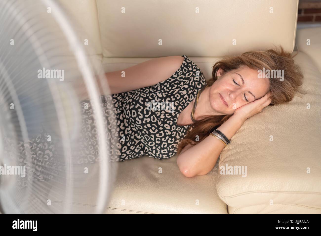 Frau, die auf dem Sofa liegt, mit einem Ventilator davor, der ihr im Sommer frische Luft gibt. Stockfoto