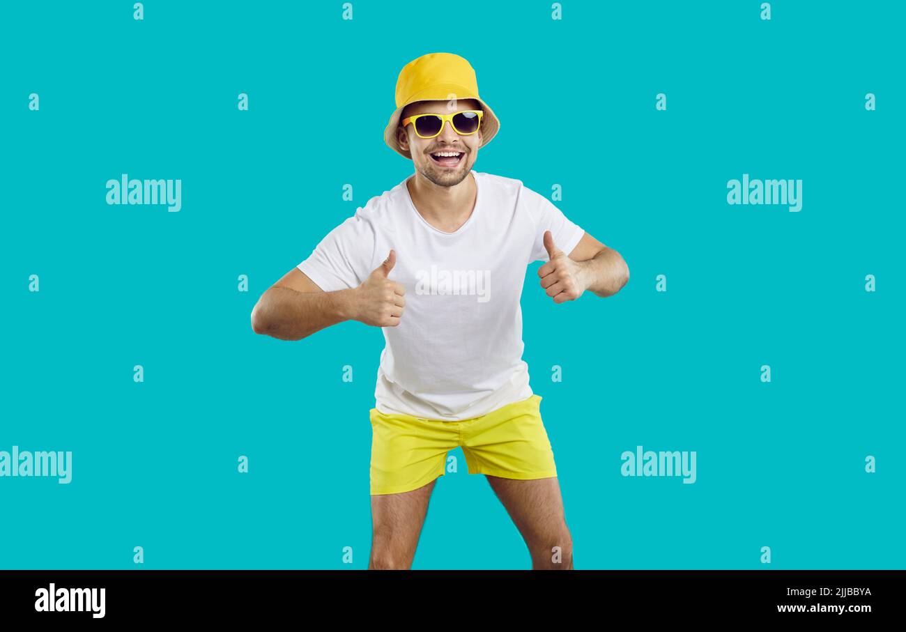 Fröhlicher, aktiver junger Mann auf hellblauem Hintergrund, der mit Daumen nach oben zeigt und Sommerurlaub empfiehlt. Stockfoto