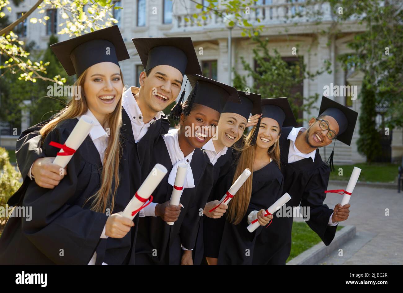 Junge, multirassische Hochschulabsolventen im akademischen Kleid posieren mit einem Lächeln nach der Abschlussfeier Stockfoto