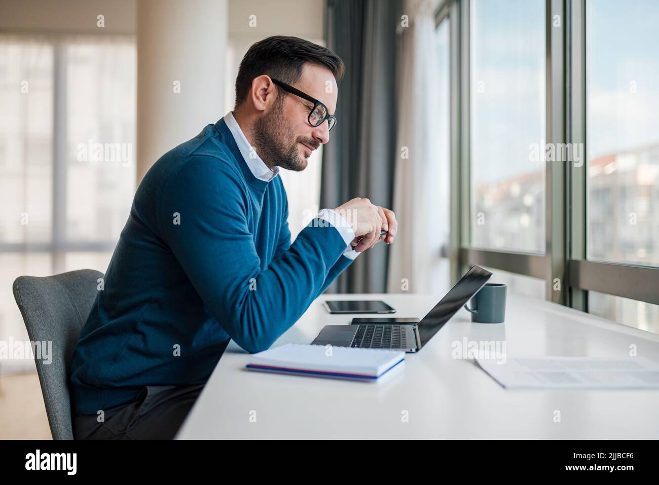 Lächelnder junger Unternehmer, der am Schreibtisch sitzt und am Laptop arbeitet. Selbstbewusster Profi analysiert erfolgreiche Strategie. Er trägt eine intelligente Casua Stockfoto