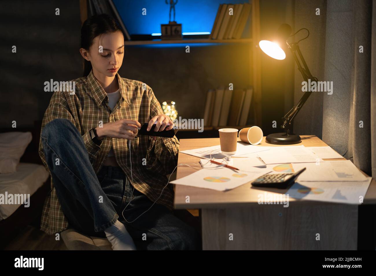 Asiatische Studentin, die nachts studiert, sich ausruht und Kopfhörer in das Telefon einsteckt, sich entspannt und Musik hört, um sich zu Hause zu entspannen Stockfoto