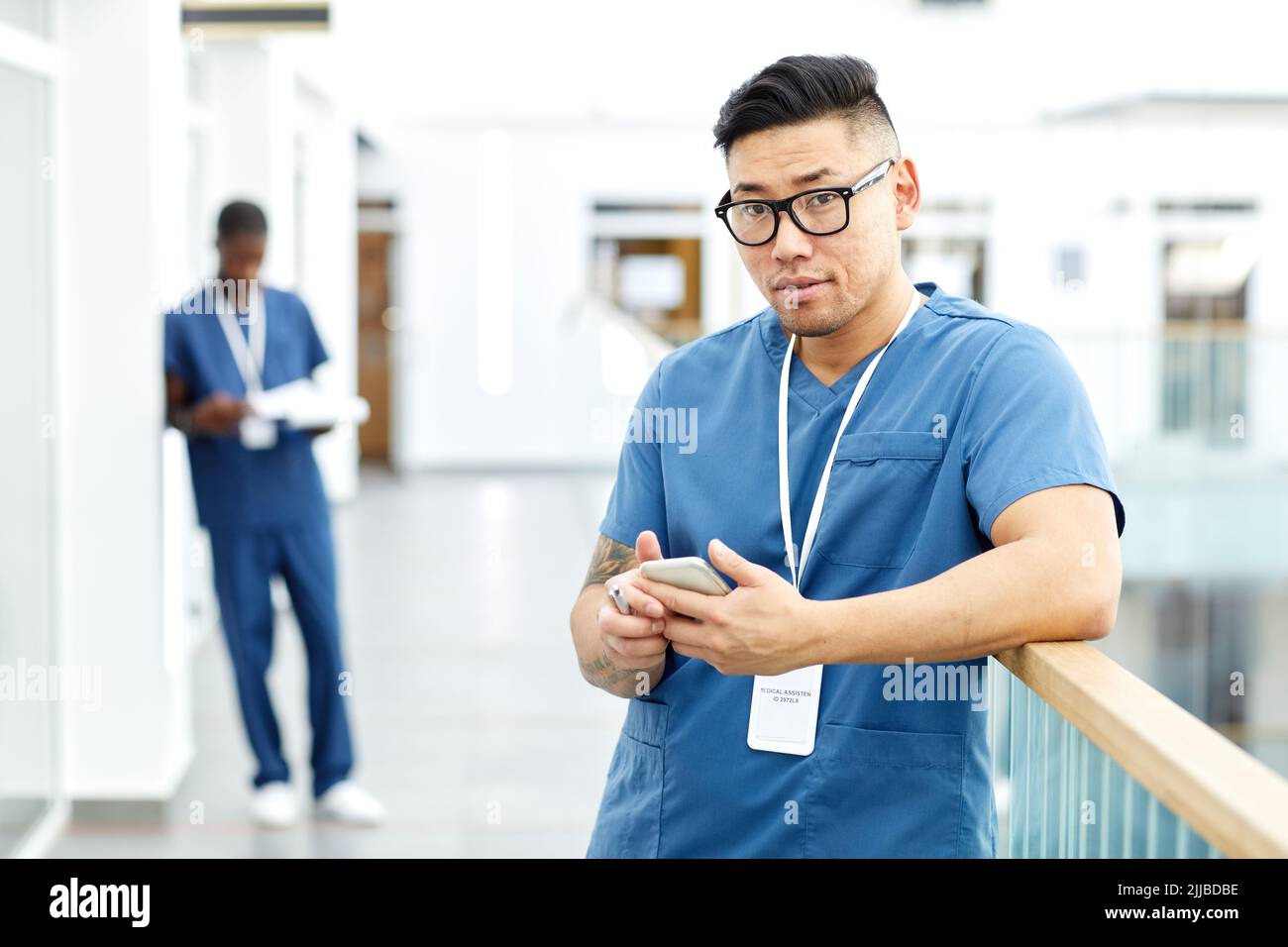 Waist-up-Porträt eines asiatischen männlichen Arztes, der die Kamera anschaut, während er das Smartphone in der Halle einer modernen Klinik benutzt, Raum kopieren Stockfoto