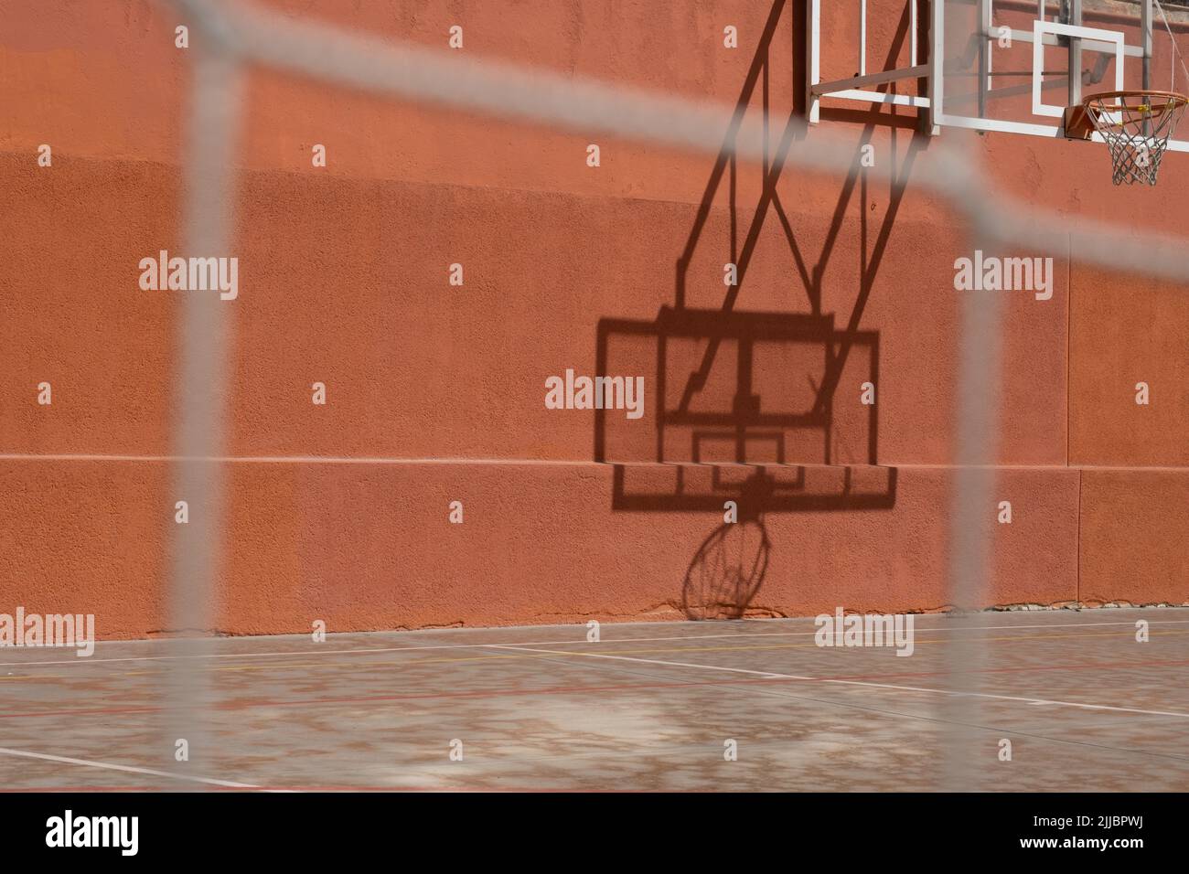 Blick aus dem niedrigen Winkel des Outdoor Basketball Backboards an einem sonnigen Tag, Schatten reflektiert auf orange Wand. Städtischer Sportplatz.verschwommene Nahaufnahme des Zauns. Stockfoto