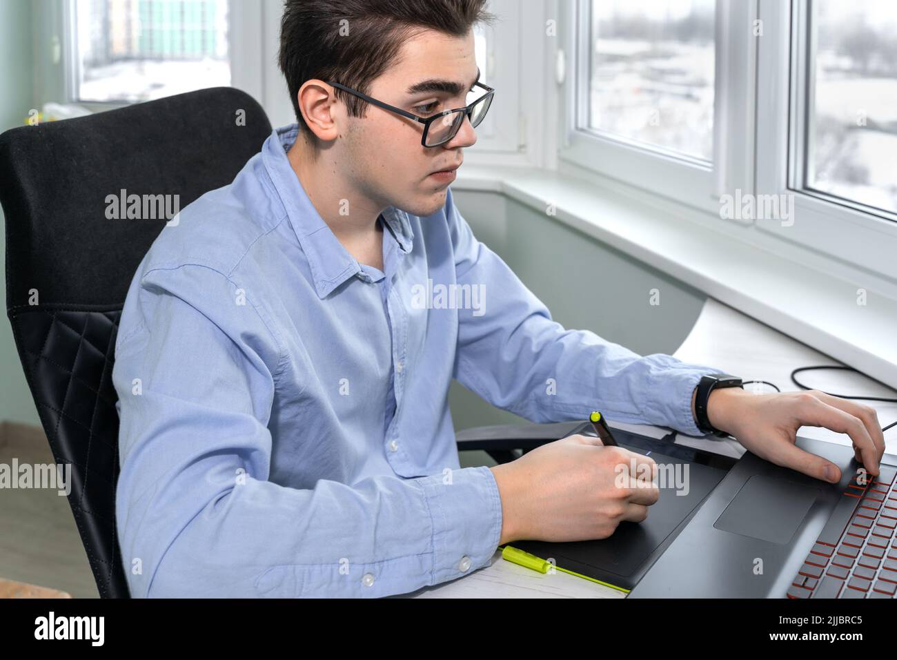 Ein junger Mann mit Brille, der mit einem Grafiktablett an seinem Laptop arbeitet Stockfoto