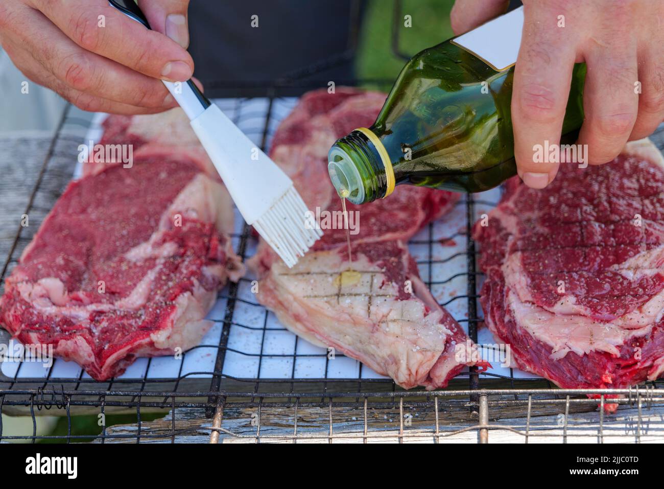 Frisches Fleisch auf Grillkorb aus Metallgeflecht. Rohes Rindersteak, ein Mann Öl das Fleisch. Leckeres Essen für den Sommer. Stockfoto