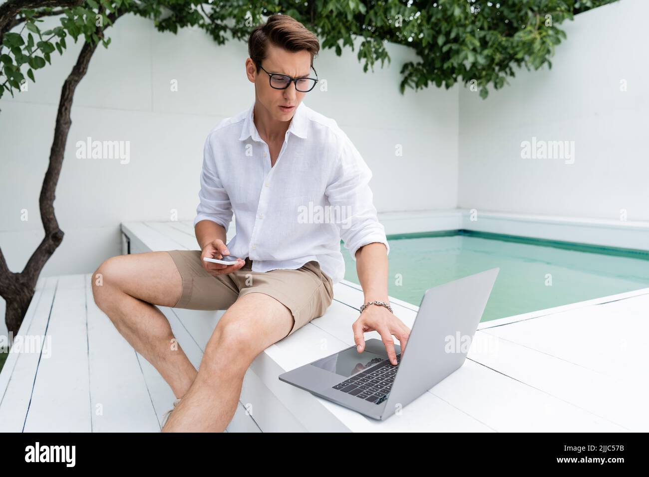 Nachdenklicher Mann in kurzen Hosen, der neben dem Pool mit Mobiltelefon sitzt und einen Laptop benutzt Stockfoto