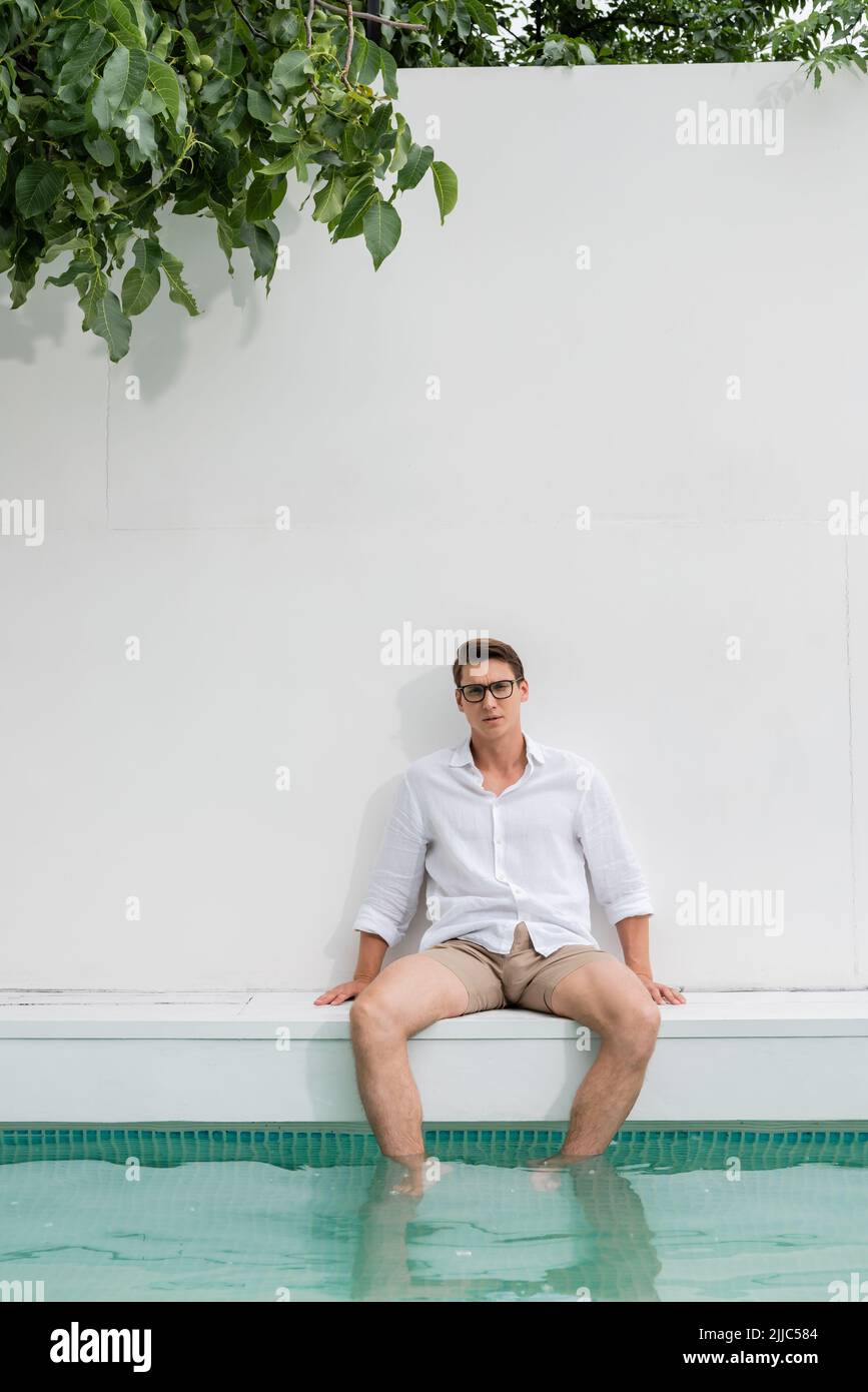 Mann in Hemd und Brille sitzt neben dem Pool an einer weißen Wand Stockfoto