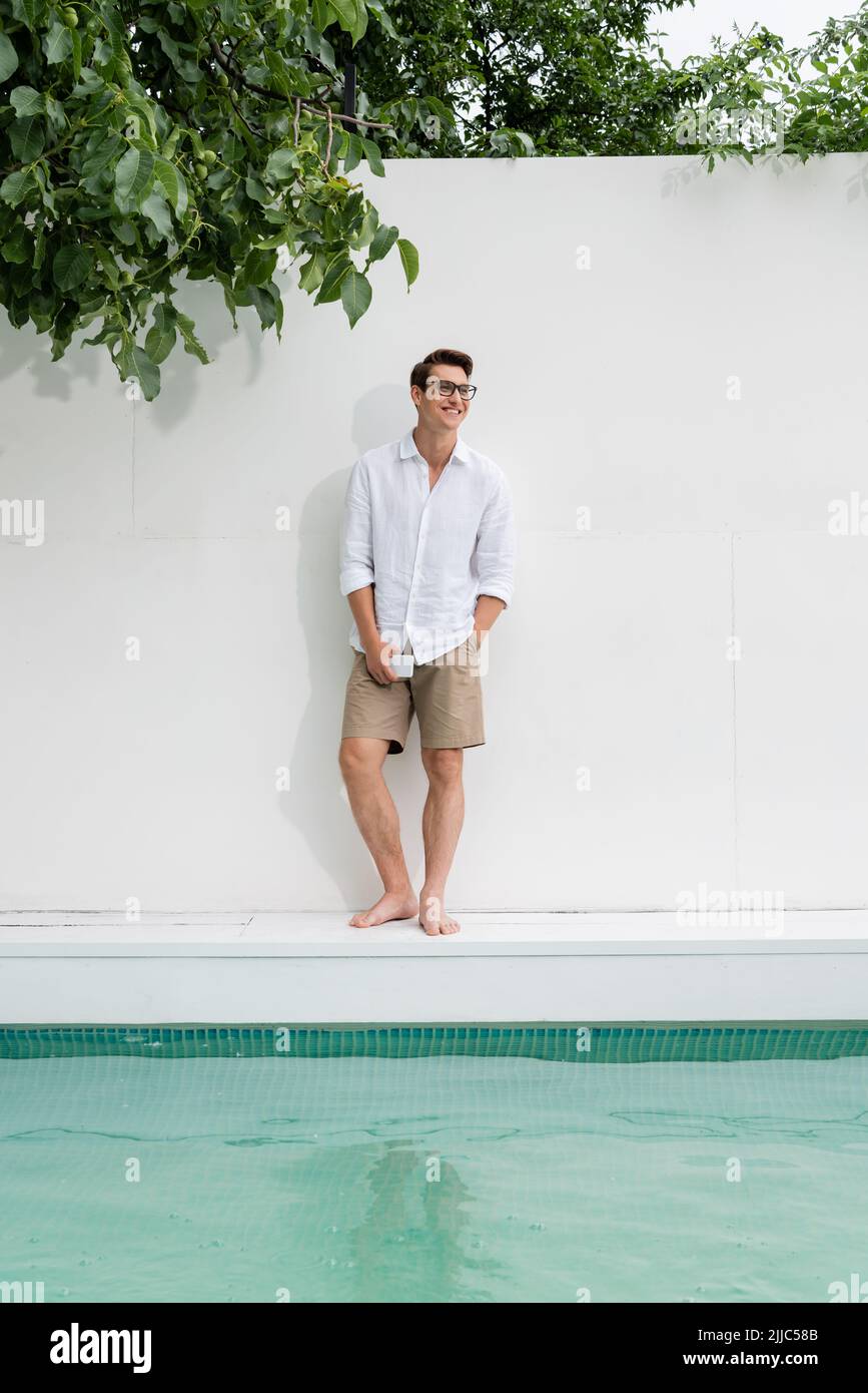 Barfuß-Mann in voller Länge, der neben dem Pool mit Smartphone und Hand in der Tasche steht Stockfoto