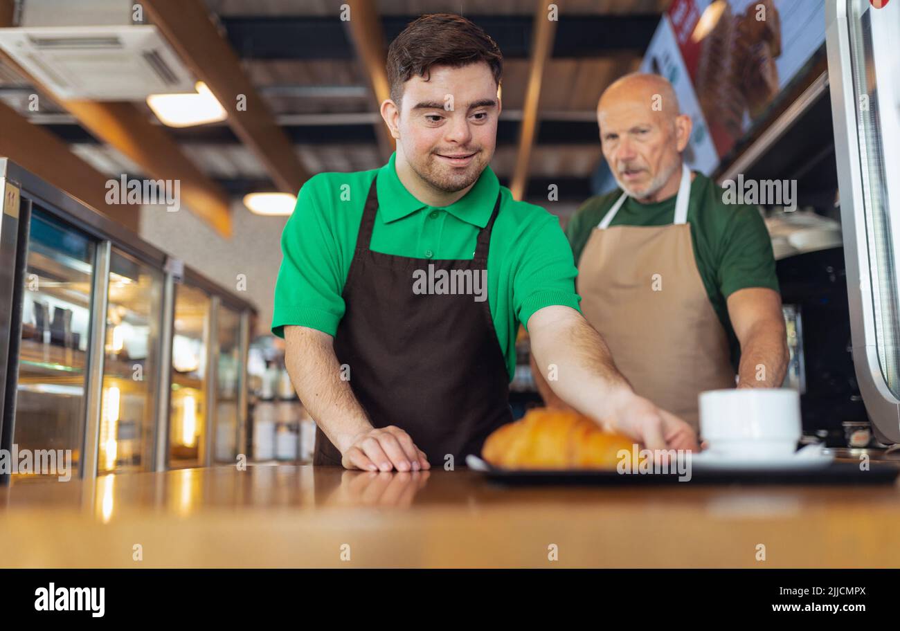 Glücklicher Kellner mit Down-Syndrom, der Kaffee mit Hilfe seines Kollegen im Café serviert. Stockfoto