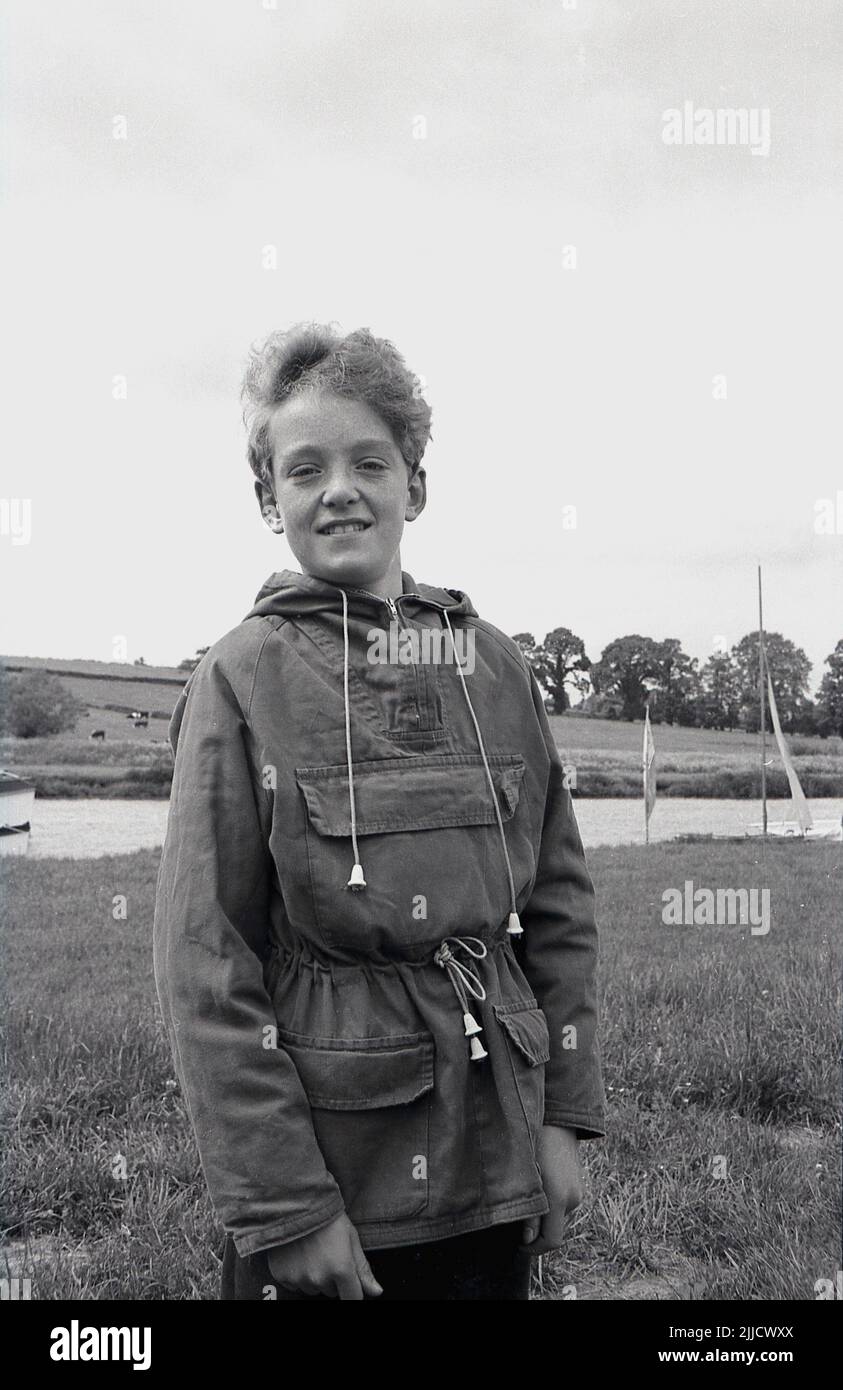 1970s, historisch, ein junger Teenager, der in der Nähe eines Flusses steht und eine Segeljacke aus Segeltuch der damaligen Zeit mit Kordelzug, Kapuze und drei Fronttaschen trägt, Tewkesbury Sailing Club, England, Großbritannien. Stockfoto