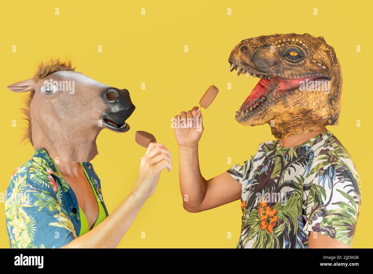 Mann in Dinosaurier Tier Kopfmaske und Frau mit Pferdekopf essen Schokolade Eis isoliert auf gelbem Hintergrund mit Kopieplatz Stockfoto