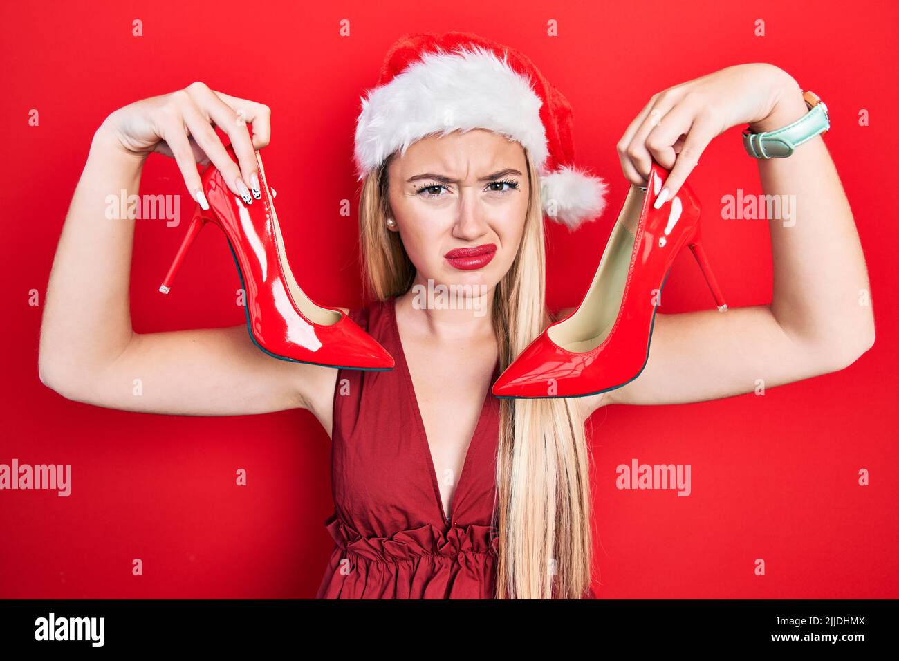 Junge blonde Mädchen trägt weihnachtshut hält High Heel Schuhe ahnungslos und verwirrt Ausdruck. Zweifel Konzept. Stockfoto