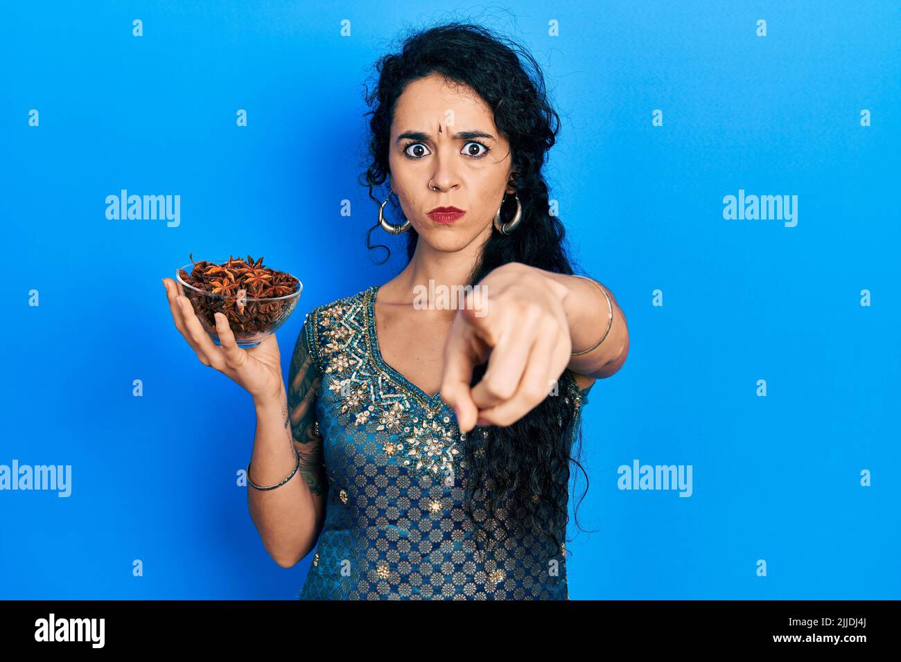 Junge Frau mit Bindi und traditionellem Kurta-Kleid, die eine Schale mit Sternanisanise hält und mit dem Finger auf die Kamera und auf Sie zeigt, selbstbewusster Gestenlook Stockfoto