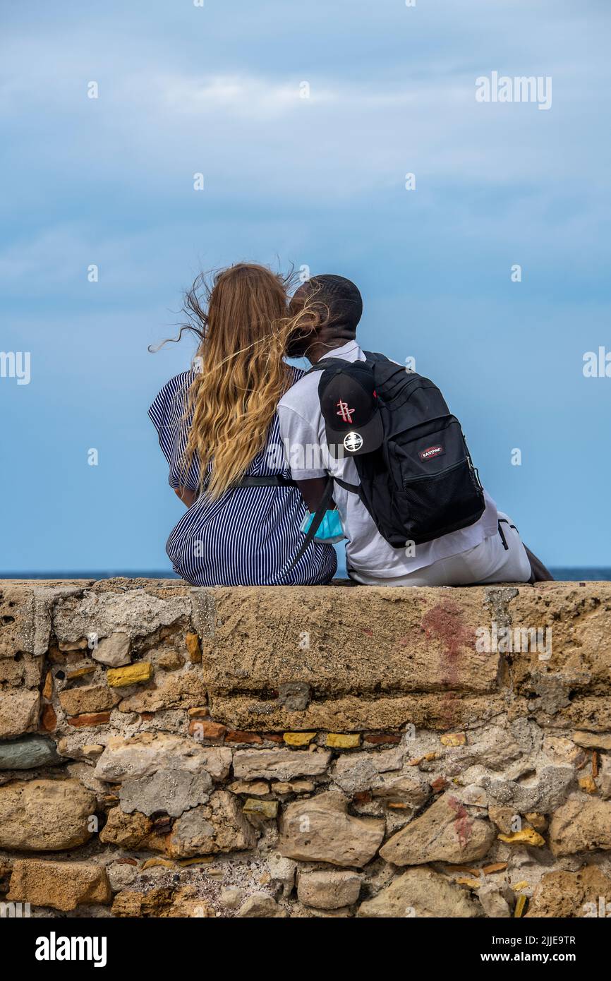 Junge Mischrenner, die an einer Wand am Meer sitzen, Mischrenner, die an einer Wand sitzen, Paare, die sich am Strand, am Uferdamm, an der Promenade umarmen, Stockfoto