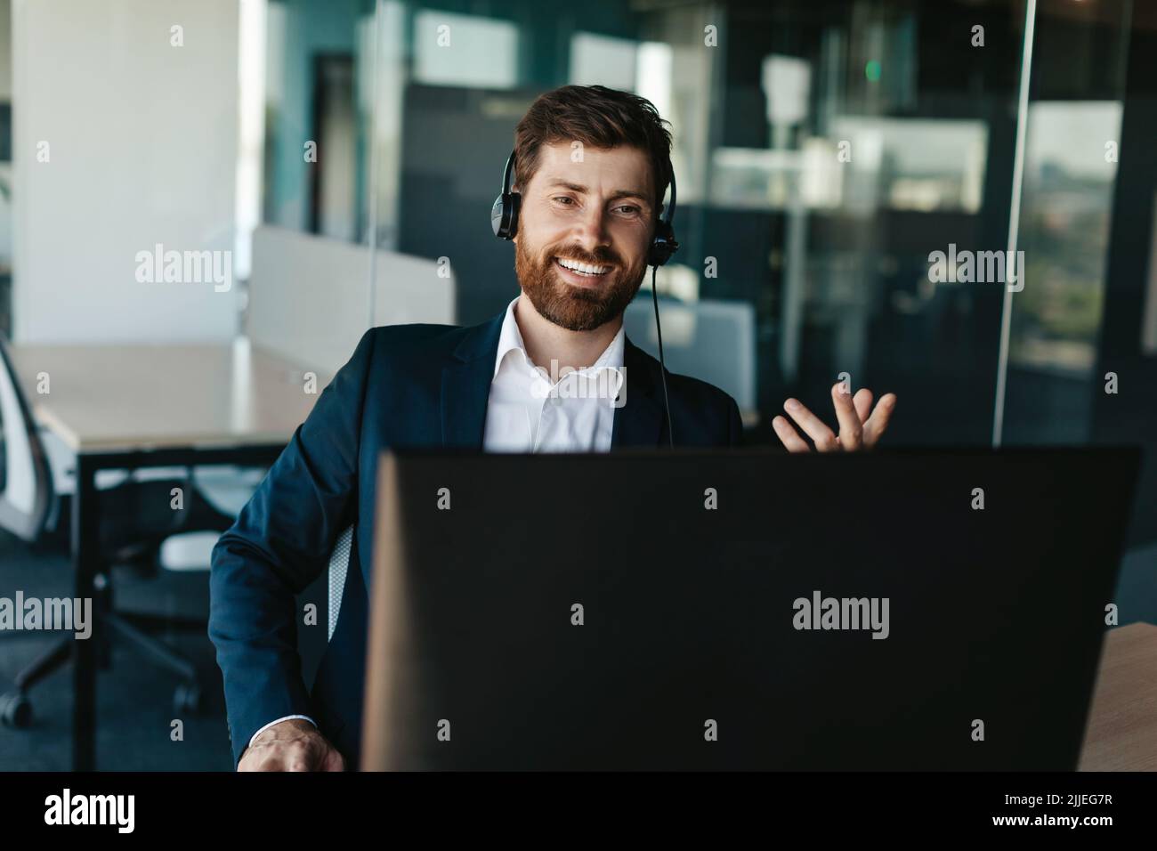 Positiver Unternehmer mit Bart im Anzug, der Kopfhörer trägt und bei der Computer-Webkamera im Büro gestikuliert Stockfoto