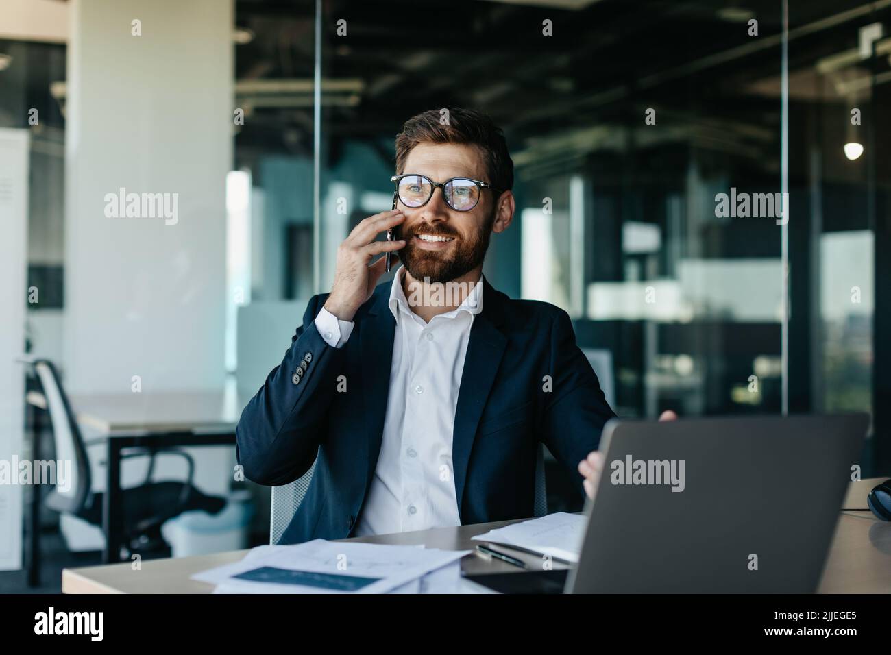 Ein glücklicher, gutaussehender Geschäftsmann, der am Laptop arbeitet und Telefongespräche führt, im Büro sitzt und lächelt Stockfoto