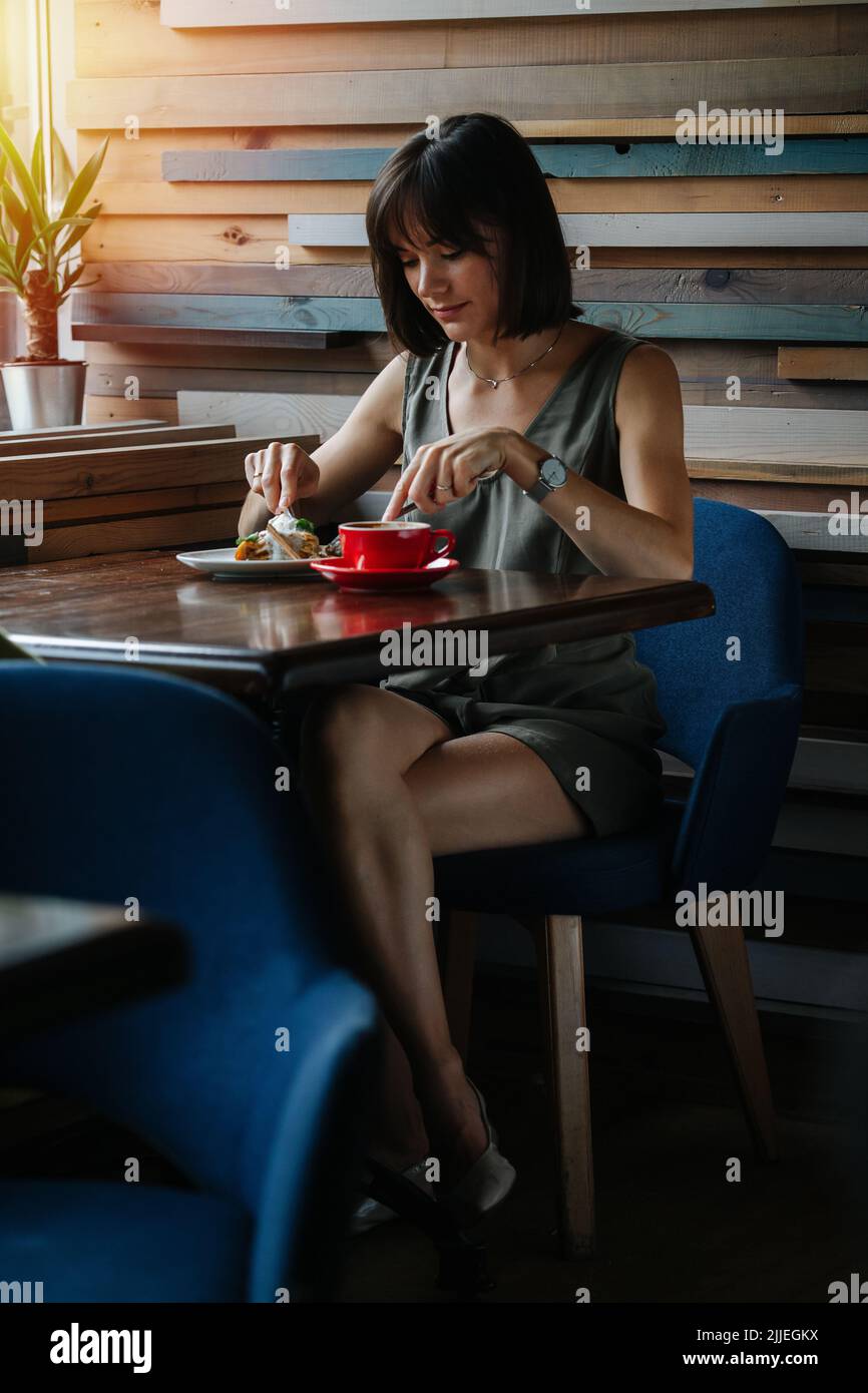 Elegante, leicht gekleidete Frau, die eine Torte mit Eiscreme und Obst mit Gabel und Messer isst. Sitzen in einer Ecke in einem Café. Stockfoto