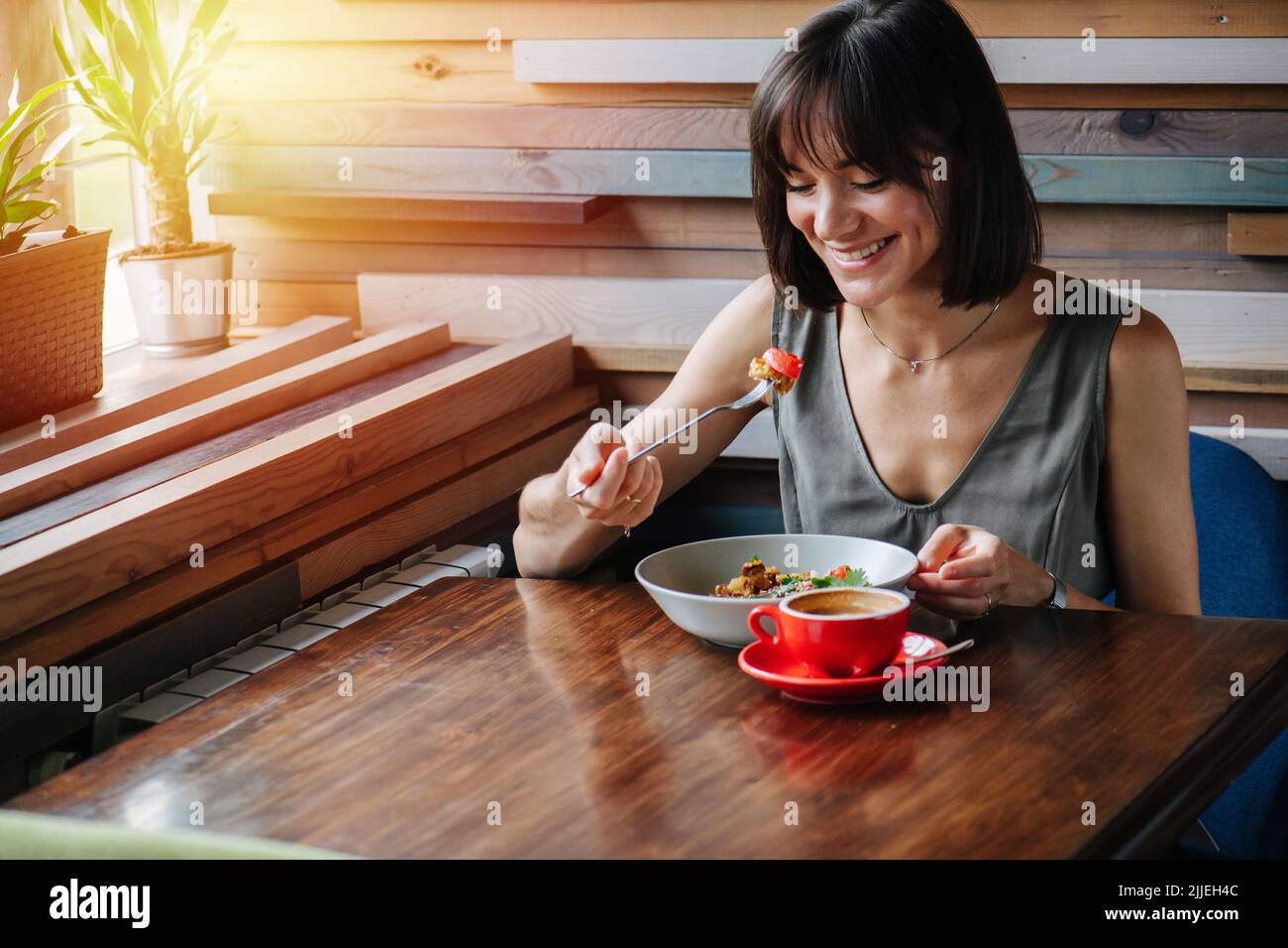 Extatische überglückliche Frau, die ein Stück Kuchen auf ihrer Gabel ansieht. Hinter einem Tisch in einem Café sitzen. Sonnenlicht, das aus einem Fenster leuchtet. Stockfoto