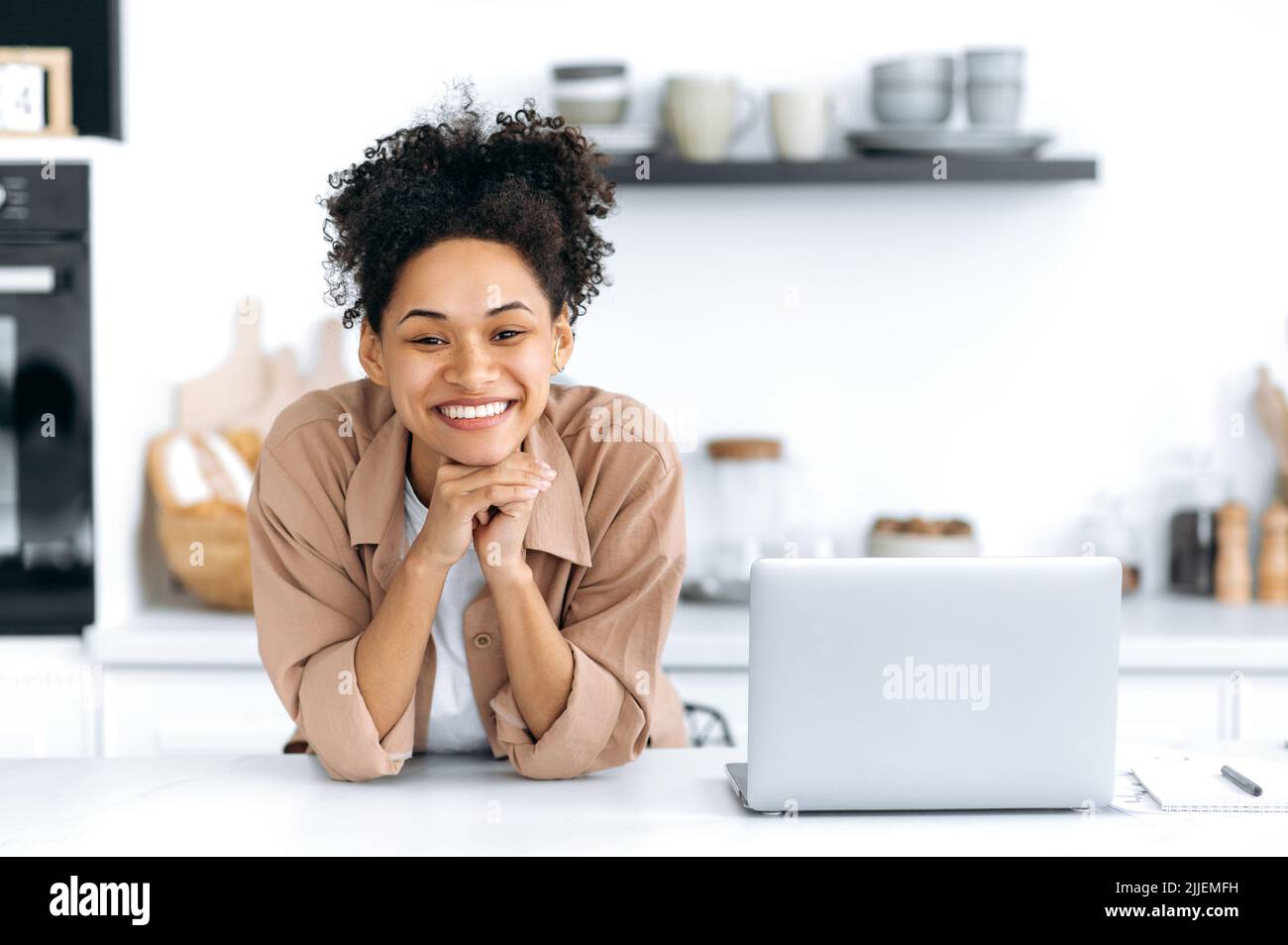 Schöne positive selbstbewusste afroamerikanische lockige junge Frau, in einem beigen Casual Shirt, steht in der Küche am Tisch neben dem Laptop, schaut auf die Kamera, lächelt glücklich Stockfoto